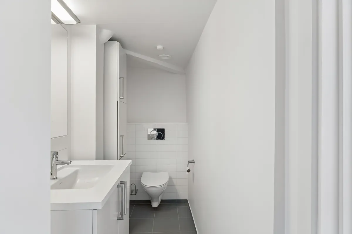 En moderne toiletrum med hvidt loft, vægge og gulv, hvidt toilet, hvidt vask og en høj hvid toiletkabinet samt hvide håndtagsgreb