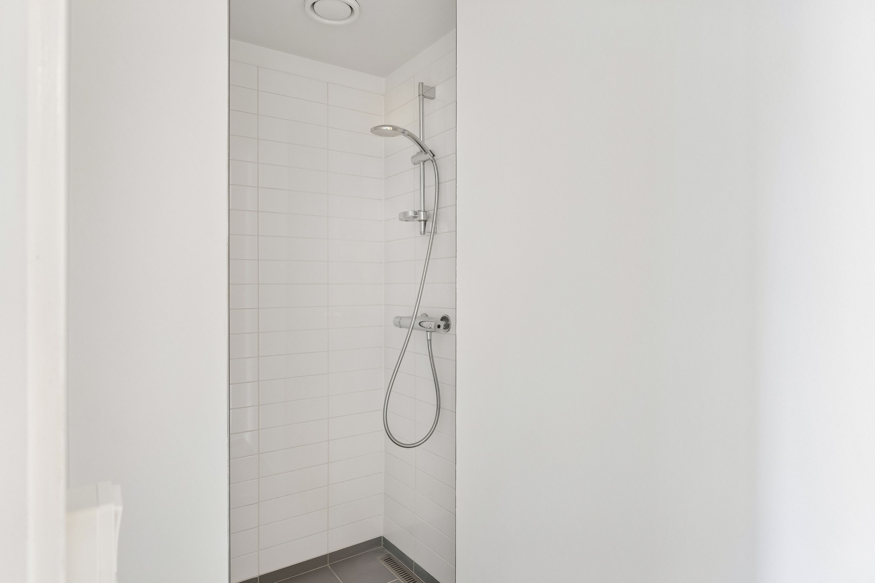 Rent og enkelt badeværelse med hvide kakeloverflader og moderne grå gulv. Simpel, sølvfarvet hånddusch med slange og vandhane. Væggene er hvide og uden særlige detaljer