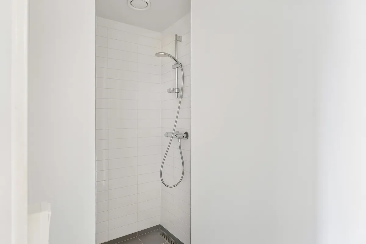 Rent og enkelt badeværelse med hvide kakeloverflader og moderne grå gulv. Simpel, sølvfarvet hånddusch med slange og vandhane. Væggene er hvide og uden særlige detaljer