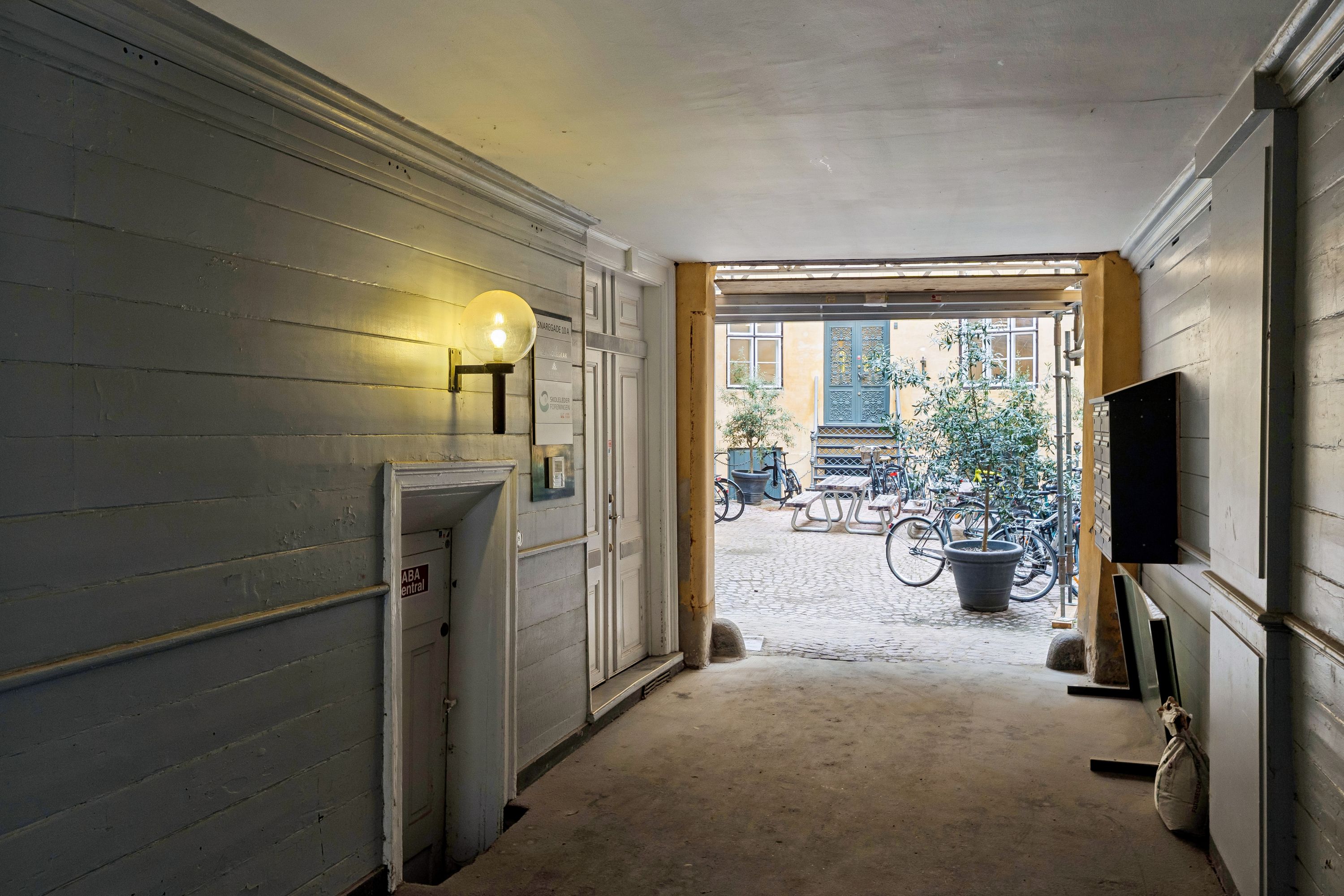 Lys pære på væg, åbne dør til gård, søjler, ruder og vinduer, blomster, skovl og skovlende søjle, træbord og stol, cykler, postkasse og postkasse og postkasse