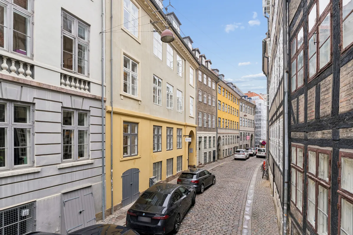 Flere biler parkeret på en lille gade med brolagte vej. En cykel står parkeret og en håndfuld biler parkeret i baggrunden. Bygninger med forskellige farvede facader og vinduer.