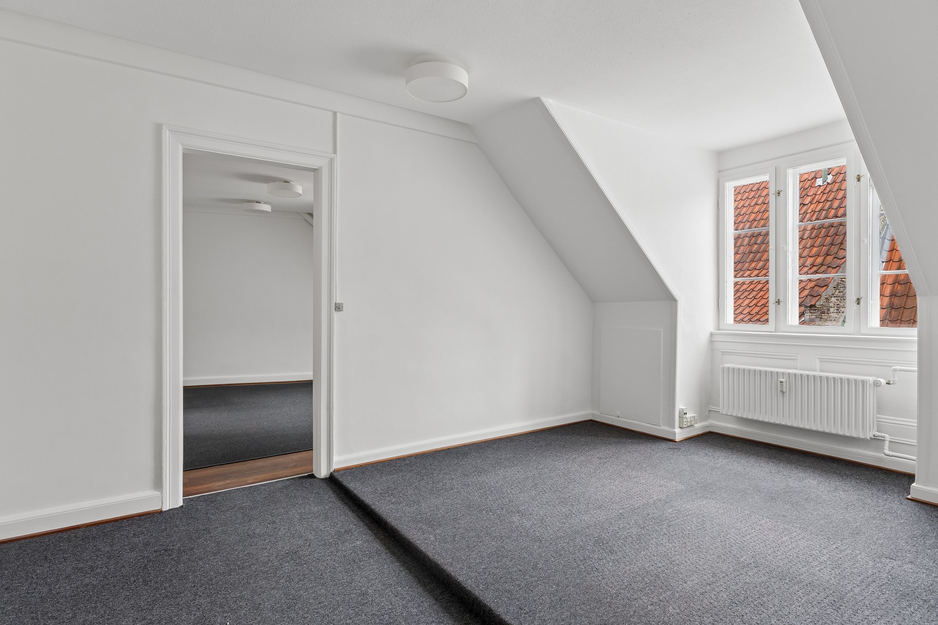 Kontor til leje i tagetagen på velholdt ejendom. Dejligt kontor med højloft, lyseruder og mørkt gulvtæppe. Lukket dør og åben vinduesåbning med røde tage