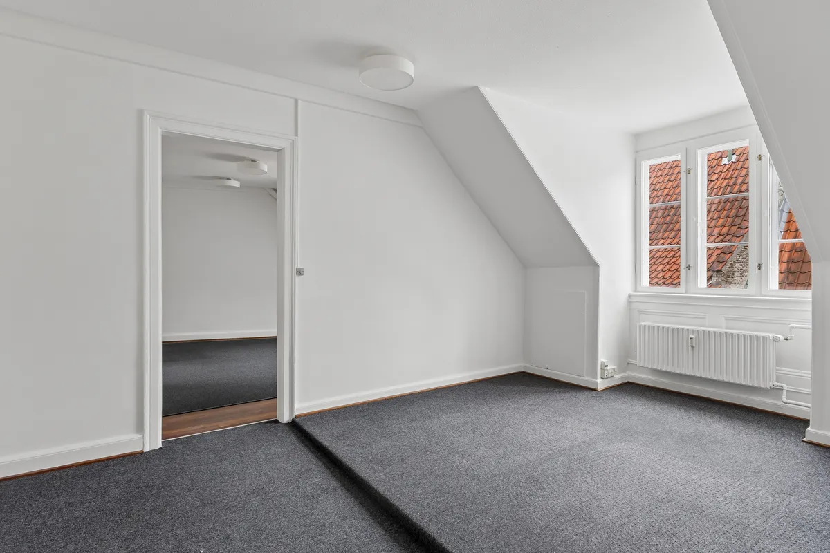 Kontor til leje i tagetagen på velholdt ejendom. Dejligt kontor med højloft, lyseruder og mørkt gulvtæppe. Lukket dør og åben vinduesåbning med røde tage