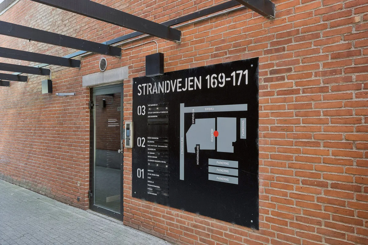 En rødmalet mur med et glasdør og et skilt med teksten Strandvejen 169-171. Et parkeringsskilt er lige under skiltet og en telefonboks er placeret ved siden af døren.