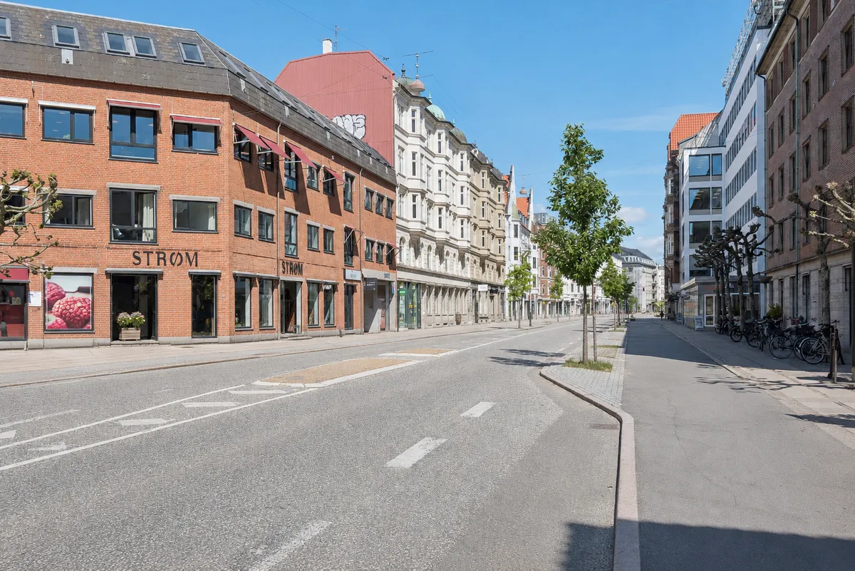 Moderne bygning med røde mursten og glasruder på Strandvejen i Hellerup. Parkerede cykler og planter på fortovet. En bred tom gadekøre og grønne træer langs gaden