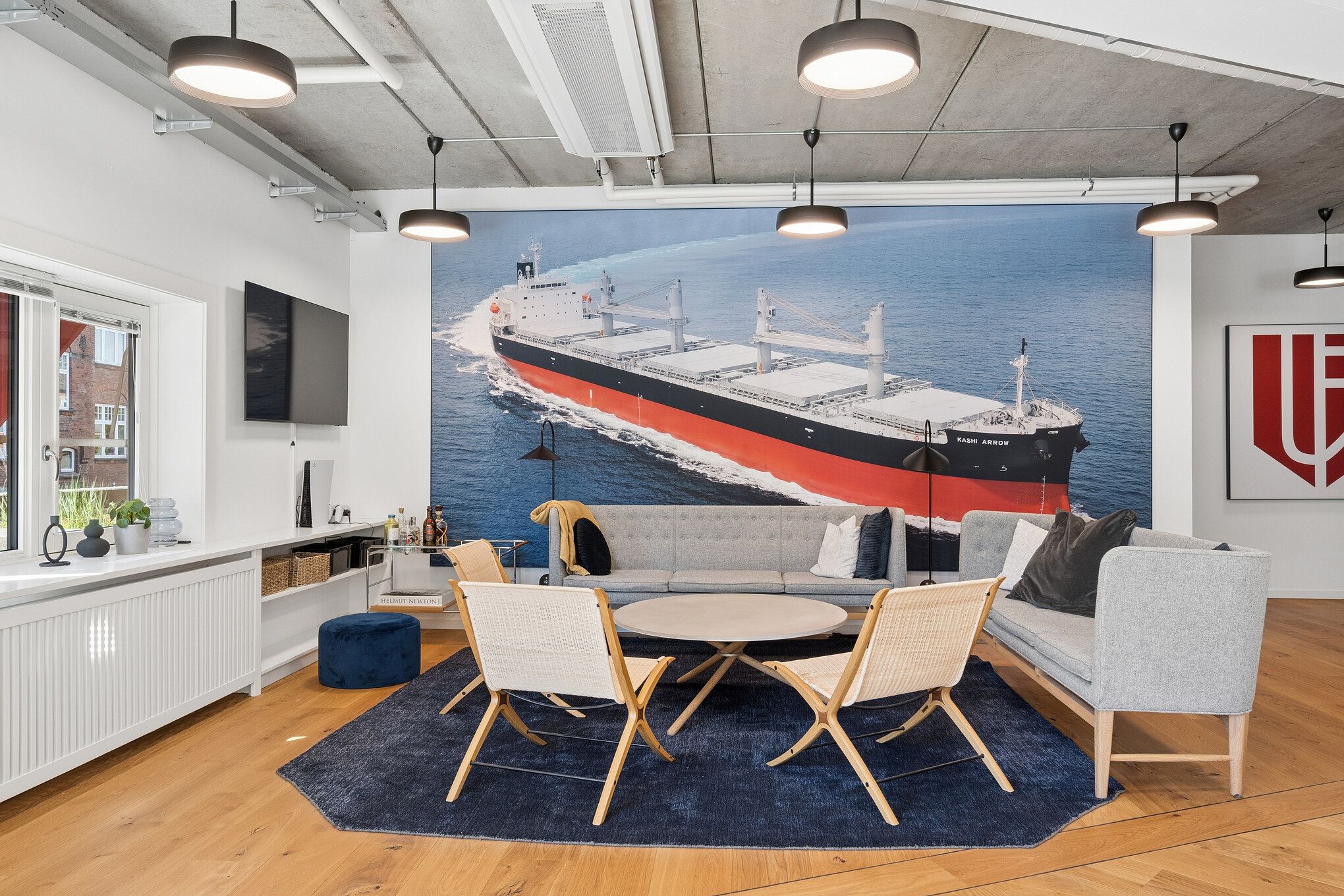 Kontorlokaler med stort skibmaleri og moderne møbler, hvid sofa, lyse lamper og vinduesudsigt over byen