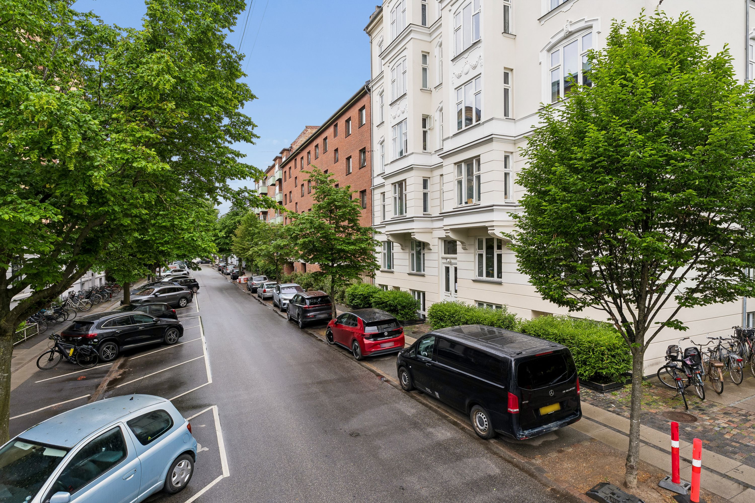 En bilparkering med mange parkerede biler, ligesom en Mercedes-Benz, Volkswagen og en bil med nummerplade. På højre side af parkeringspladsen er der et hus med mange vinduer.