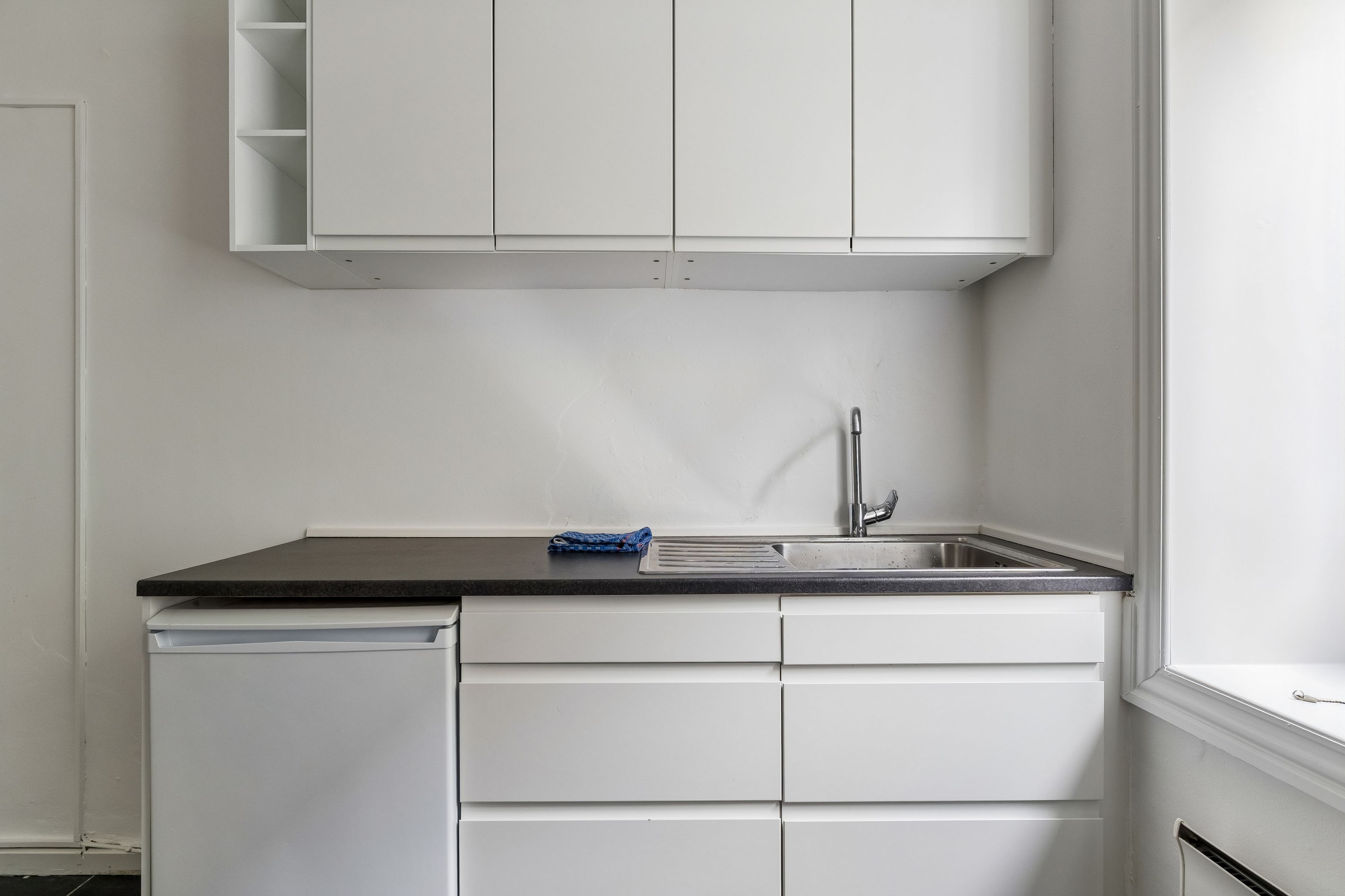 Kontor med lyse loftskappe, hvidt køkken med lysegråt bord, hvidt køleskab og sort køkkenbord, samt høj, smal vinduesåbning med lysets refleksion på gulvet