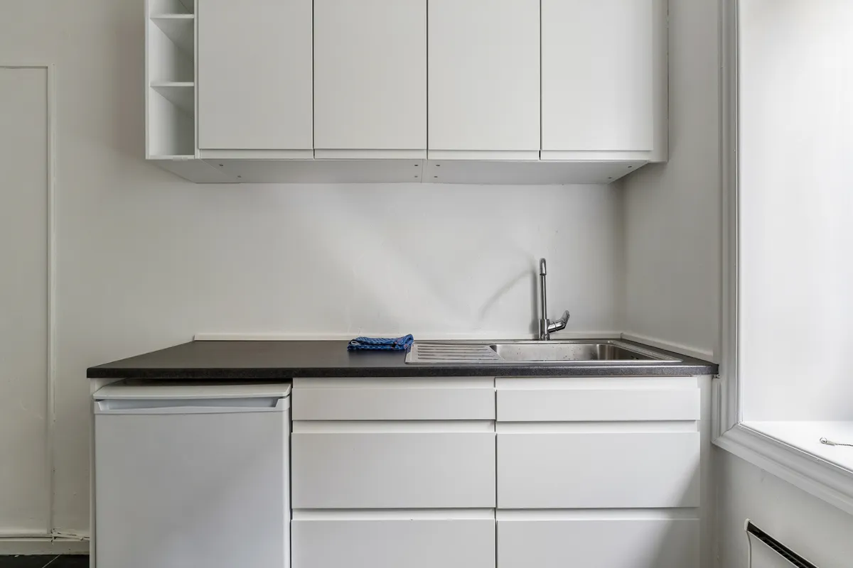 Kontor med lyse loftskappe, hvidt køkken med lysegråt bord, hvidt køleskab og sort køkkenbord, samt høj, smal vinduesåbning med lysets refleksion på gulvet
