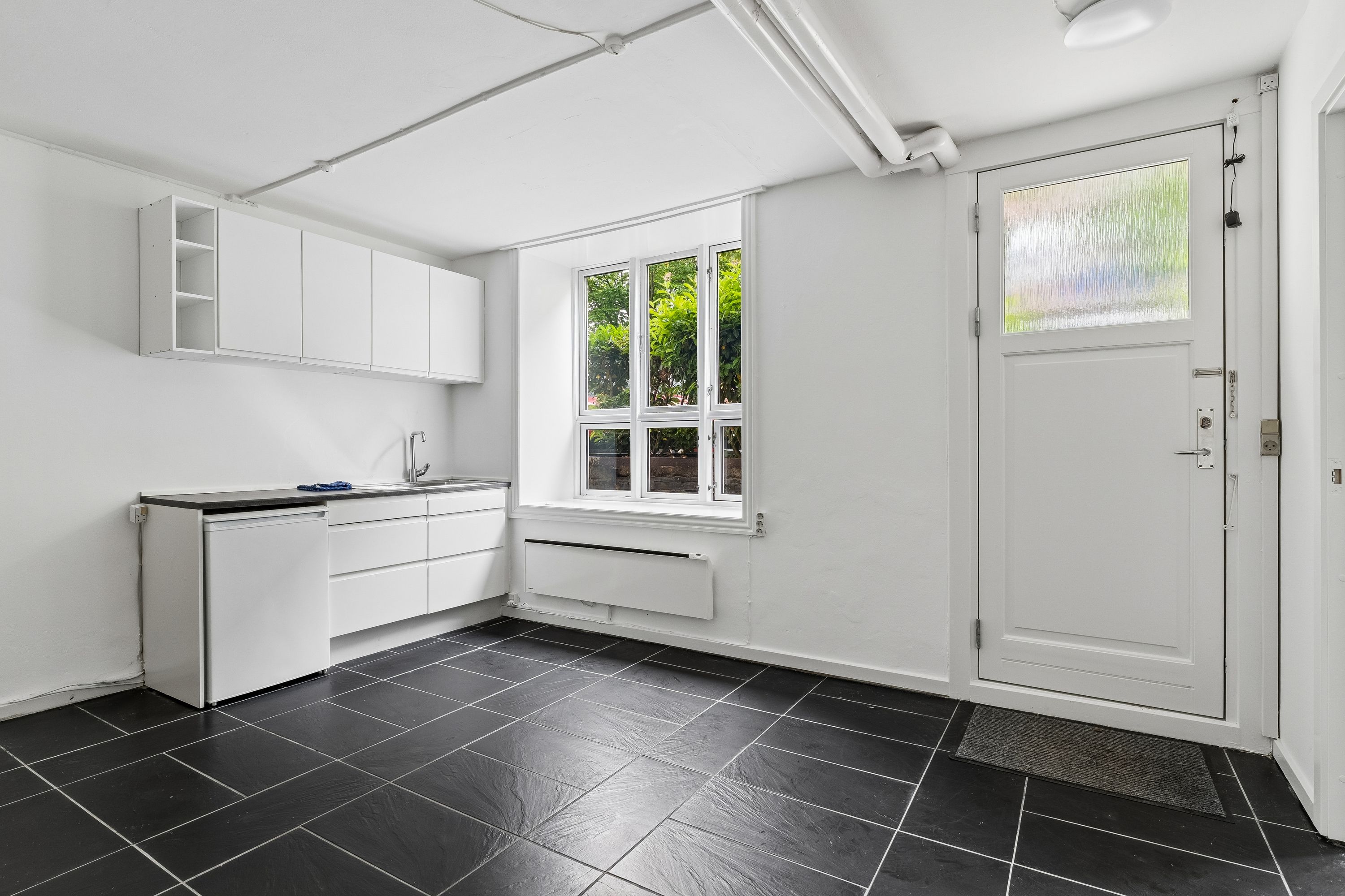 En minimalistisk køkken med hvide vægge og sort fliser. Hvidt køkken med køleskab, vask og hængende hylder. Et vindue med udsigt til grønne planter og vej
