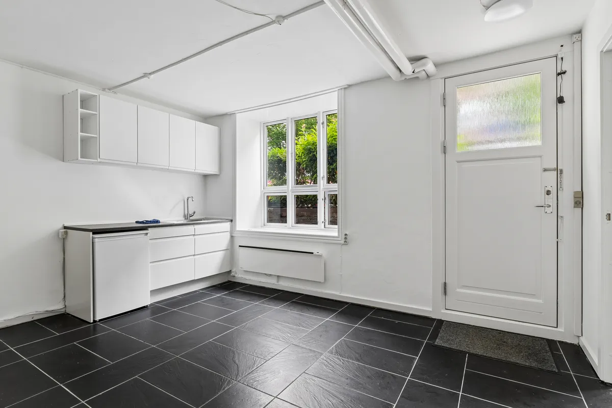 En minimalistisk køkken med hvide vægge og sort fliser. Hvidt køkken med køleskab, vask og hængende hylder. Et vindue med udsigt til grønne planter og vej