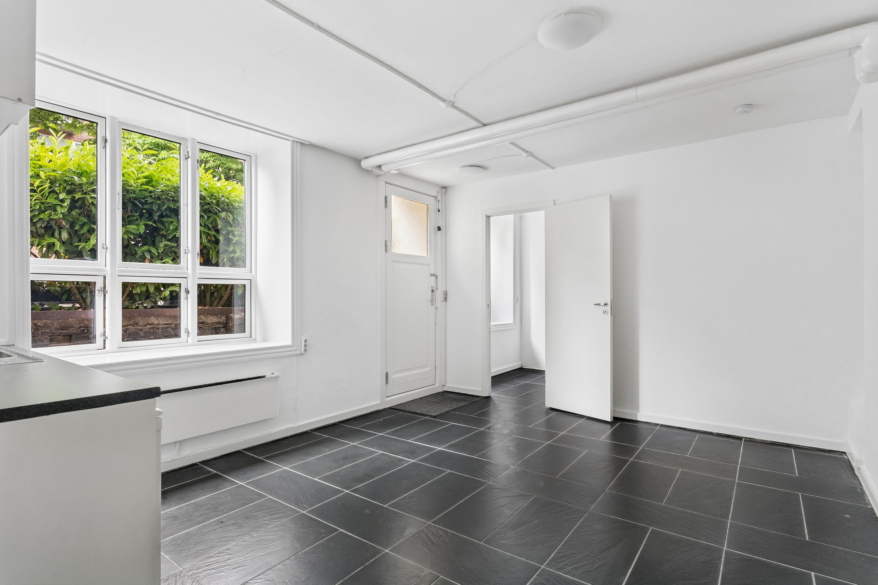 Lej kontorlejlighed på Harsdorffsvej 7, Frederiksberg. 35 m² med hvidt interiør og smukke sort gulv. Vindue med udsigt til buske og sten. Hvidt køkken og moderne design.