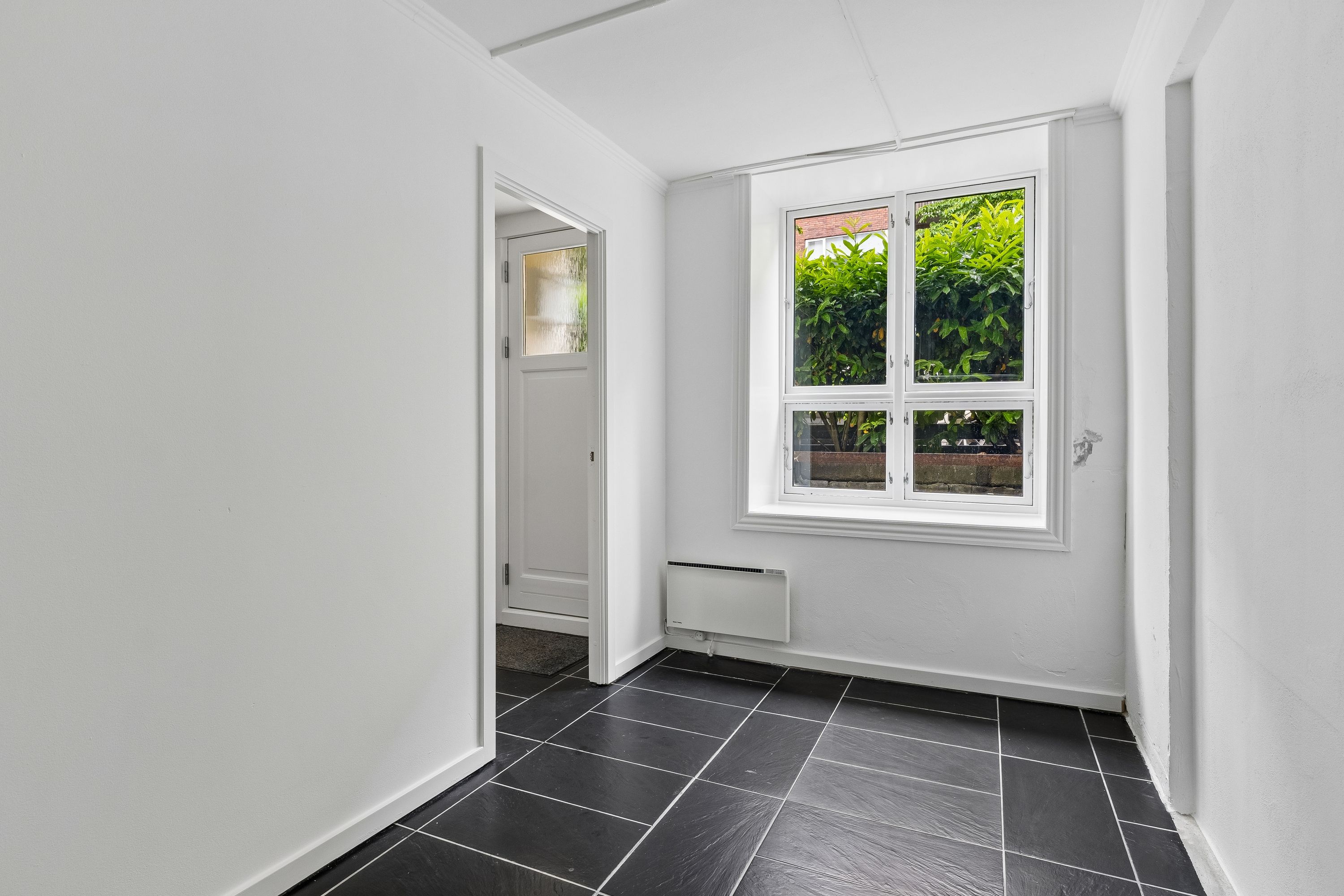 Sorte flisegulv og hvide vægge i kontorlokaler med et vindue mod haven og en hvid dør. Et hvidt radiator er monteret på væggen.