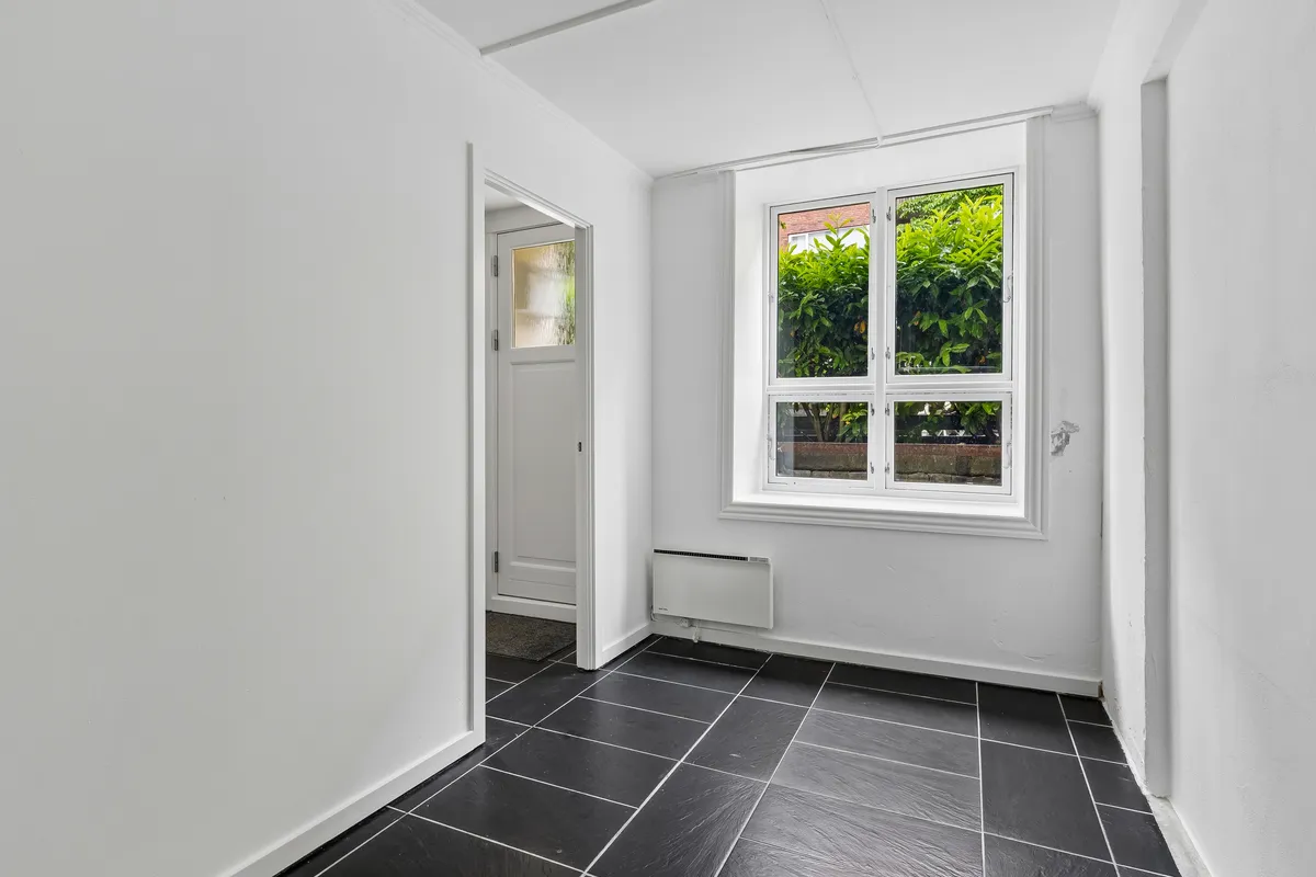 Sorte flisegulv og hvide vægge i kontorlokaler med et vindue mod haven og en hvid dør. Et hvidt radiator er monteret på væggen.