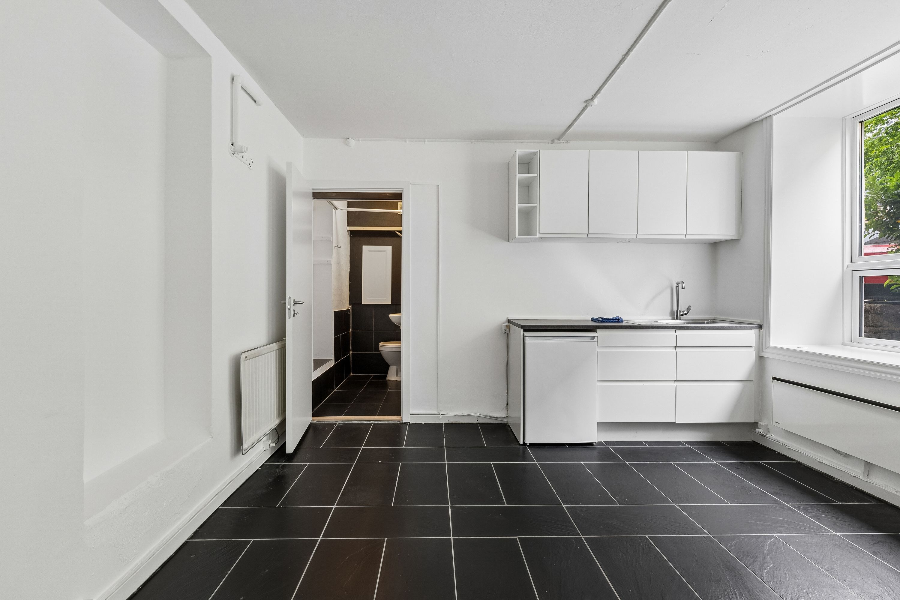 Værelse med hvidt loft og væg og sort flisegulv. Rent og funktionelt med køkken og toilet. Moderne hvide skabe og disk.