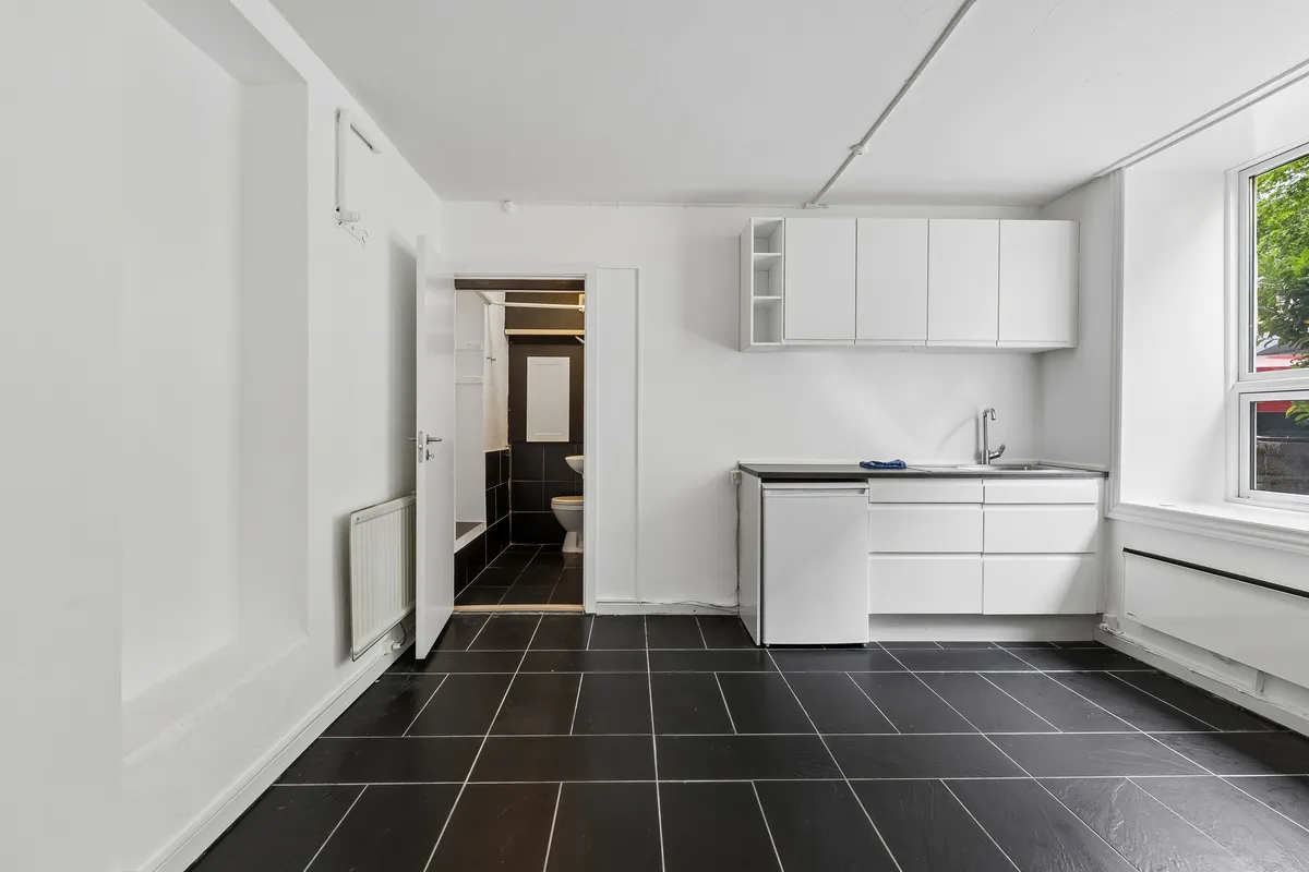 Værelse med hvidt loft og væg og sort flisegulv. Rent og funktionelt med køkken og toilet. Moderne hvide skabe og disk.