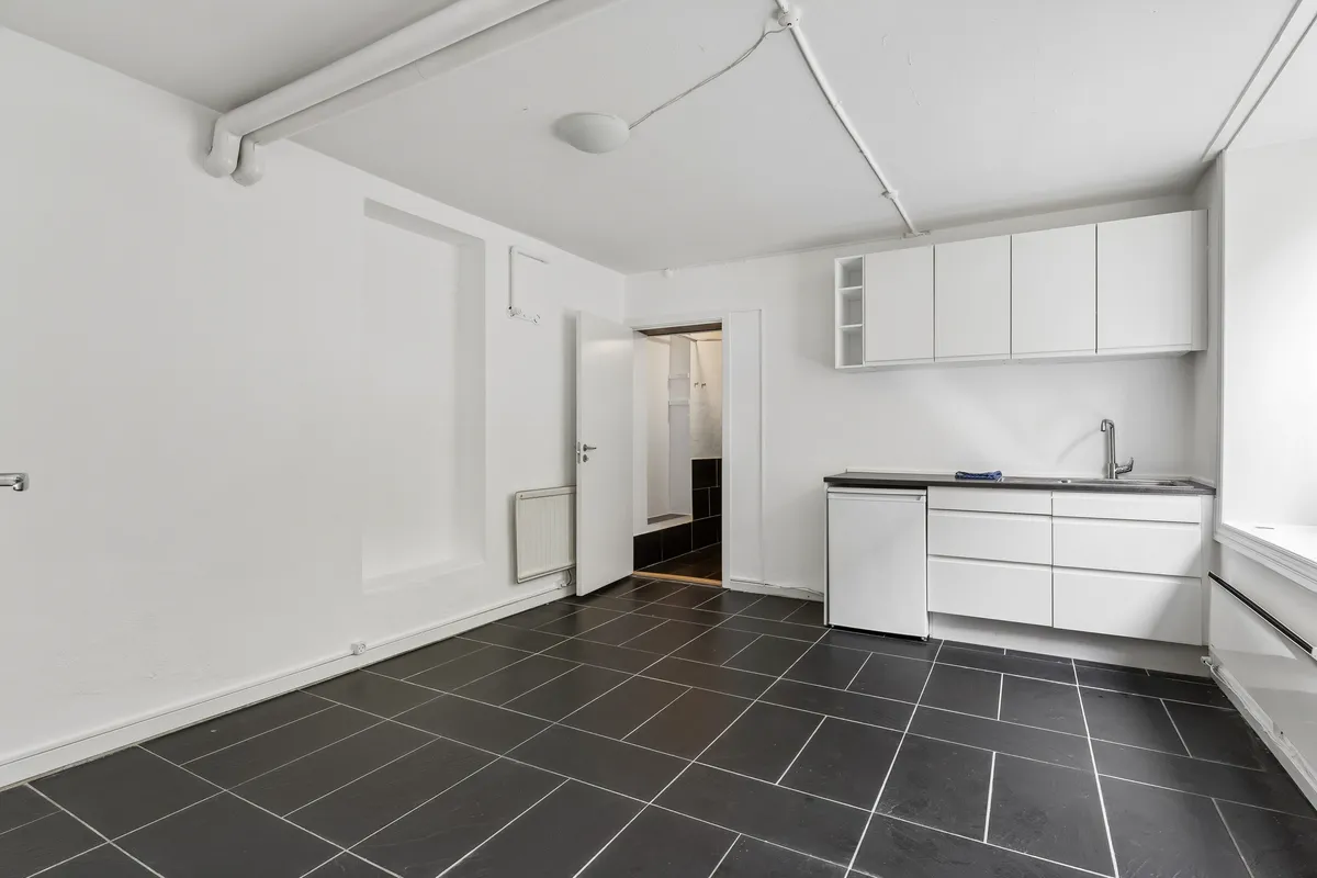 Rent og moderne kontorrum med hvid væg og sortgule gulvfliser. Der er hvide kabinetter, hvide skabe, en hvid komfur og en hvid radiator. Der er et hvidt loft med en hvid ventil og en hvid lampe.