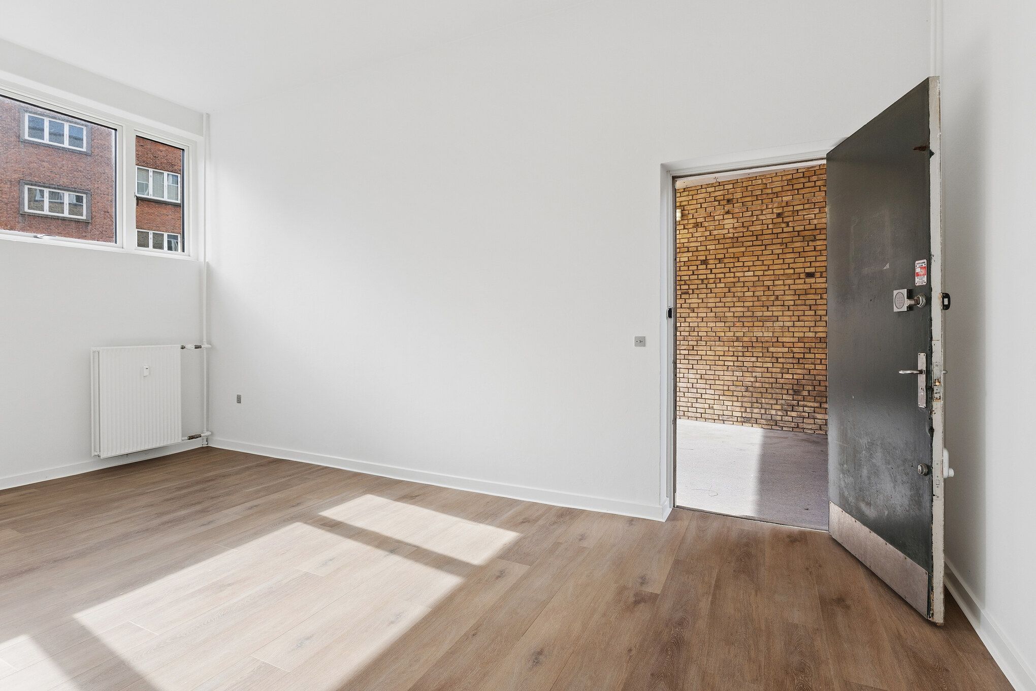En tom, lyse, hvidrummet med trægulv og åben dør, hvor gulvet skinner og reflekterer sollys. Et hvidt radiator er placeret på væggen.