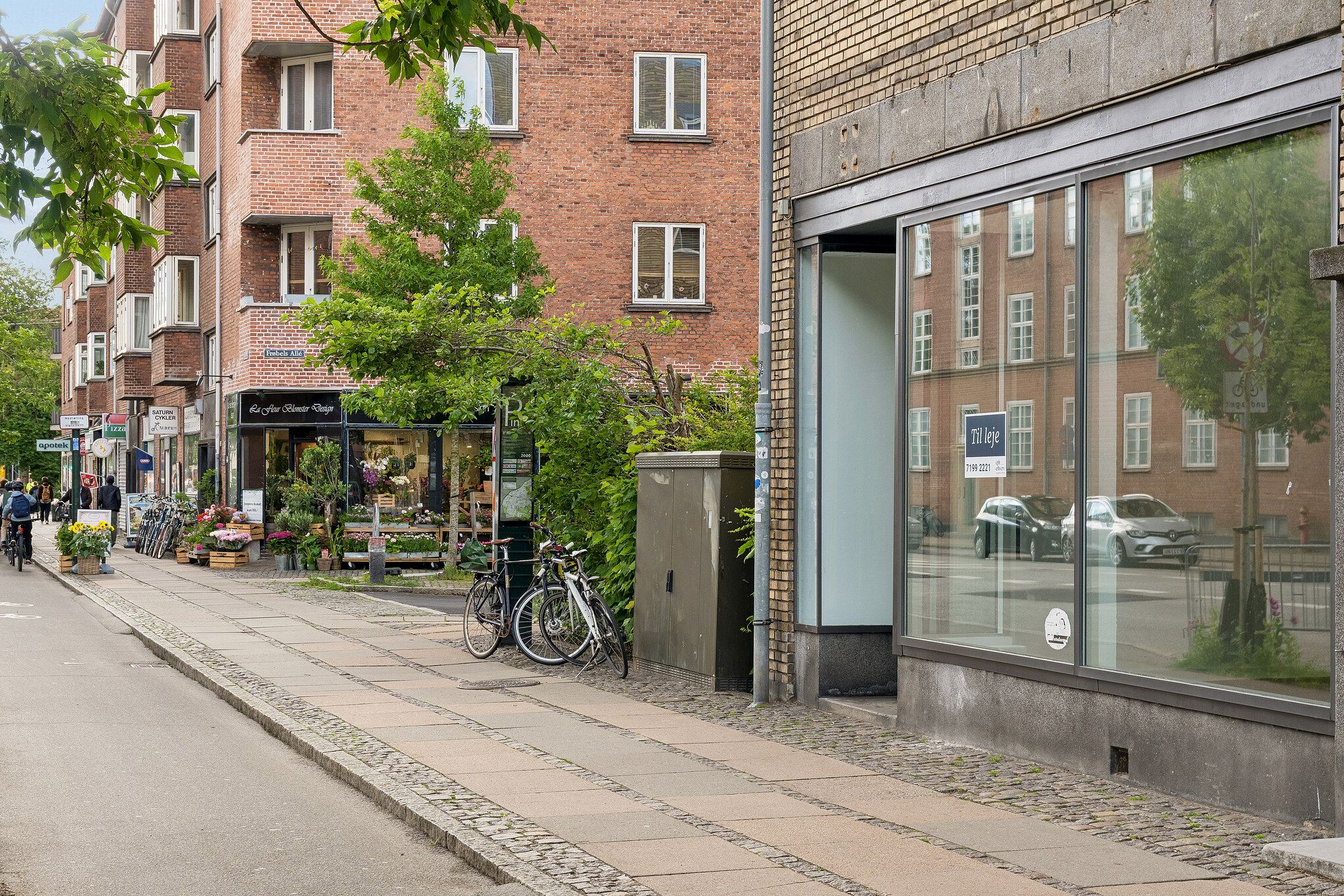 Store butikker og blomsterforretning på Finsensvej i Frederiksberg, med cyklister, grønne planter og glasvinduer mod vejen med biler og bygninger