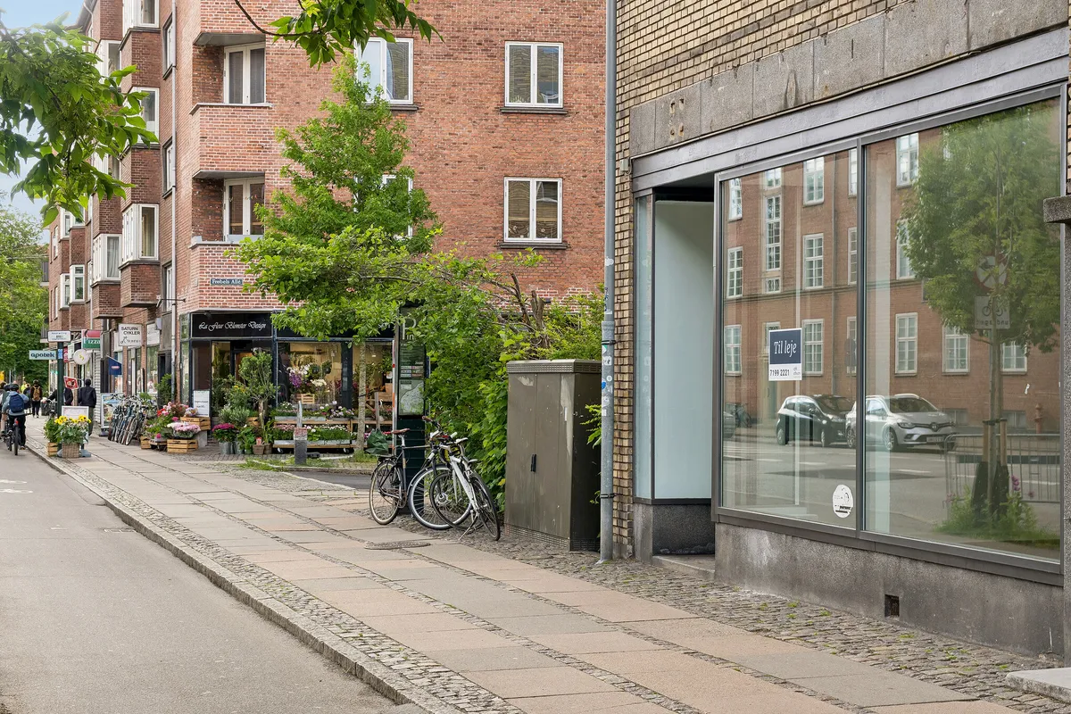 Store butikker og blomsterforretning på Finsensvej i Frederiksberg, med cyklister, grønne planter og glasvinduer mod vejen med biler og bygninger