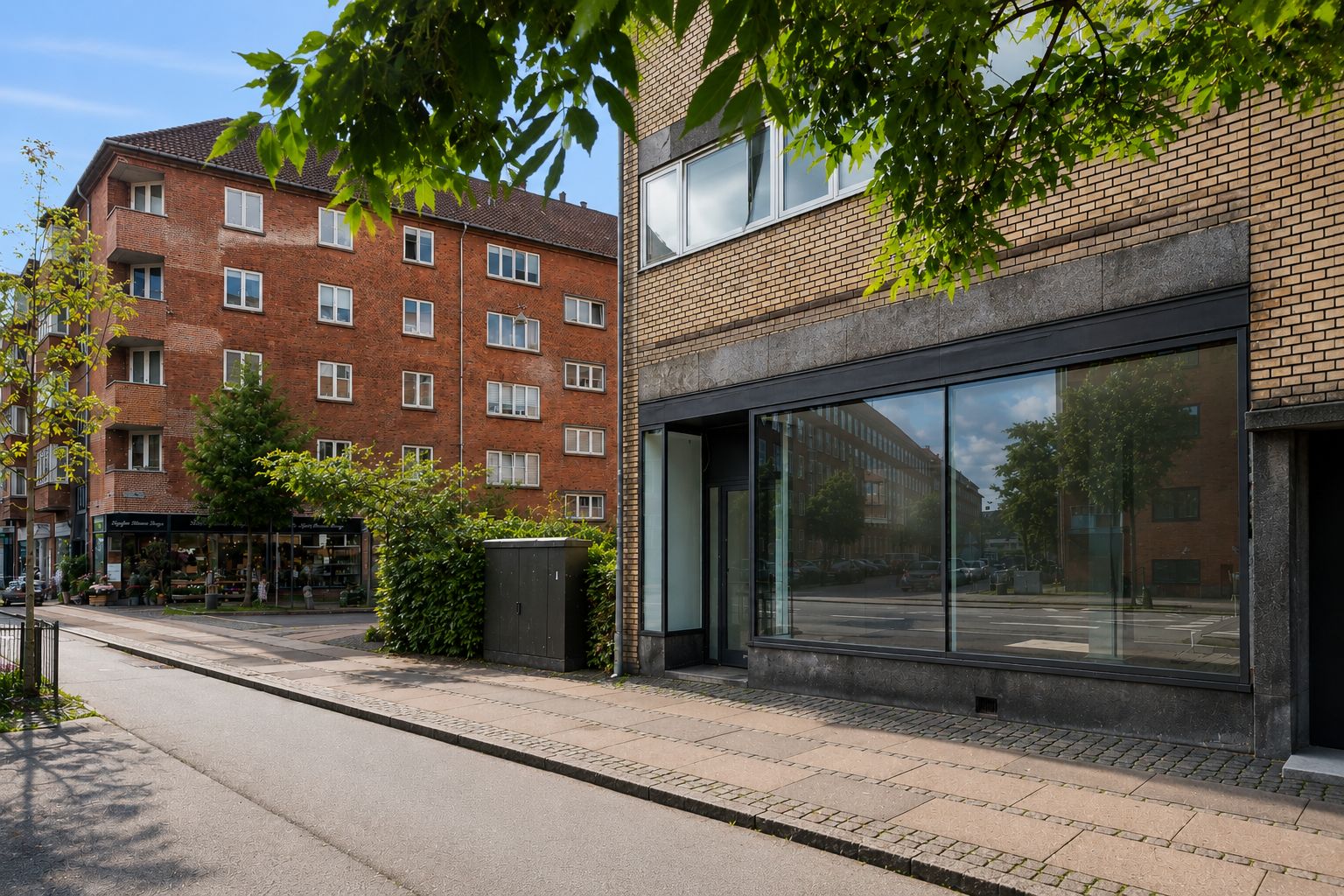 Butikslejemål i Frederiksberg. Butik med store ruder og glasdøre. En bygning med røde mursten og vinduer. Planter og grønne træer foran bygningen. Et hus med røde mursten og vinduer i baggrunden.