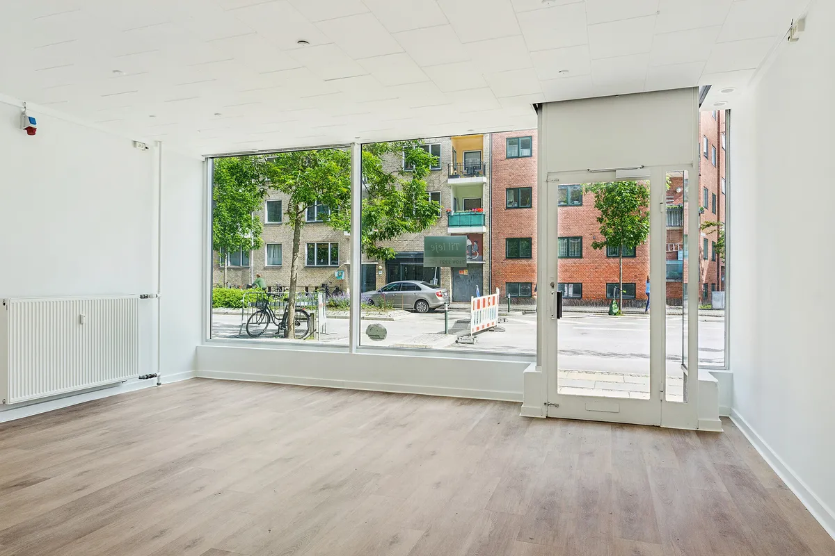 Butikslokaler med store vinduer og højt loft. Moderne interiør med lysegulve og hvide vægge. Udsigt til biler og grønne træer på gågaden.