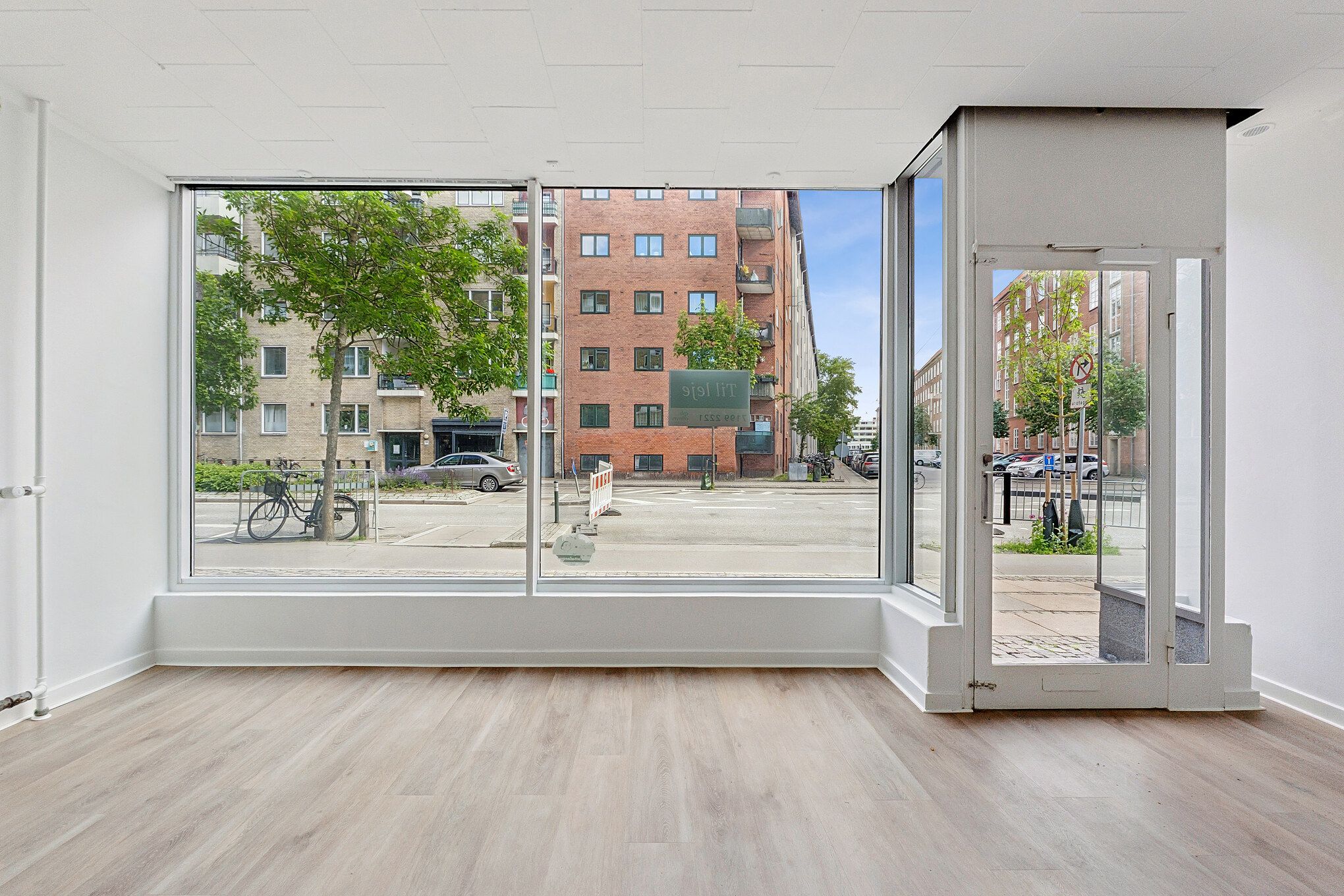 Store vinduer og højloft med hårdt trægulv i lejlighed i første sal. Udenfor er biler, cykler og grønne træer. En rødmalet bygning med høj vinduer og en grøn buske.