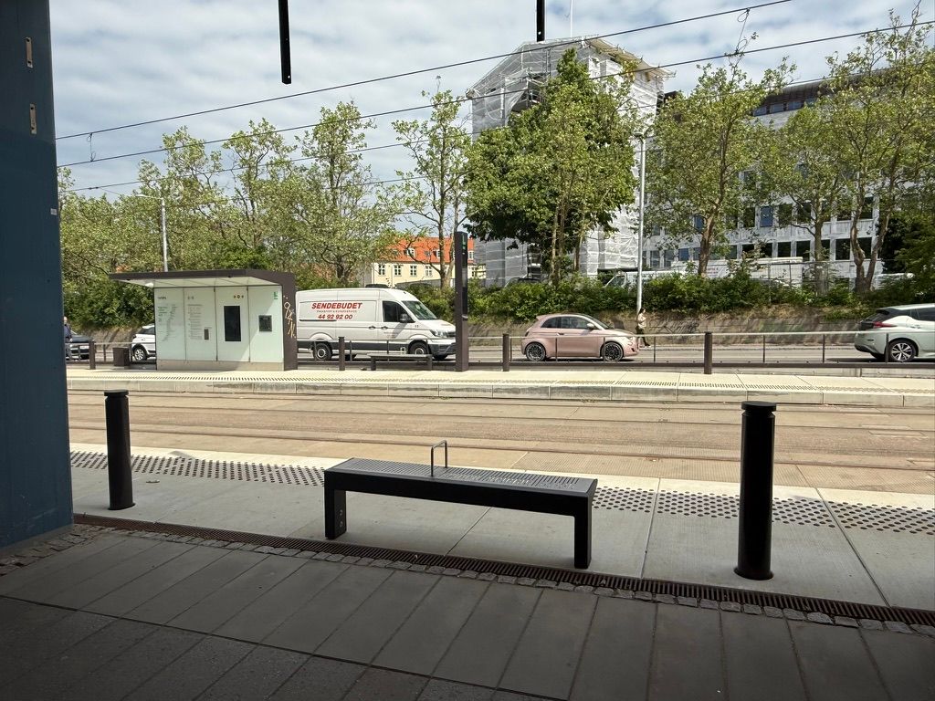 Take-away til leje på Jernbanepladsen 61 i Kongens Lyngby med parkerede biler og et busstop. I baggrunden er der bygninger og trær og en person går over gågaden