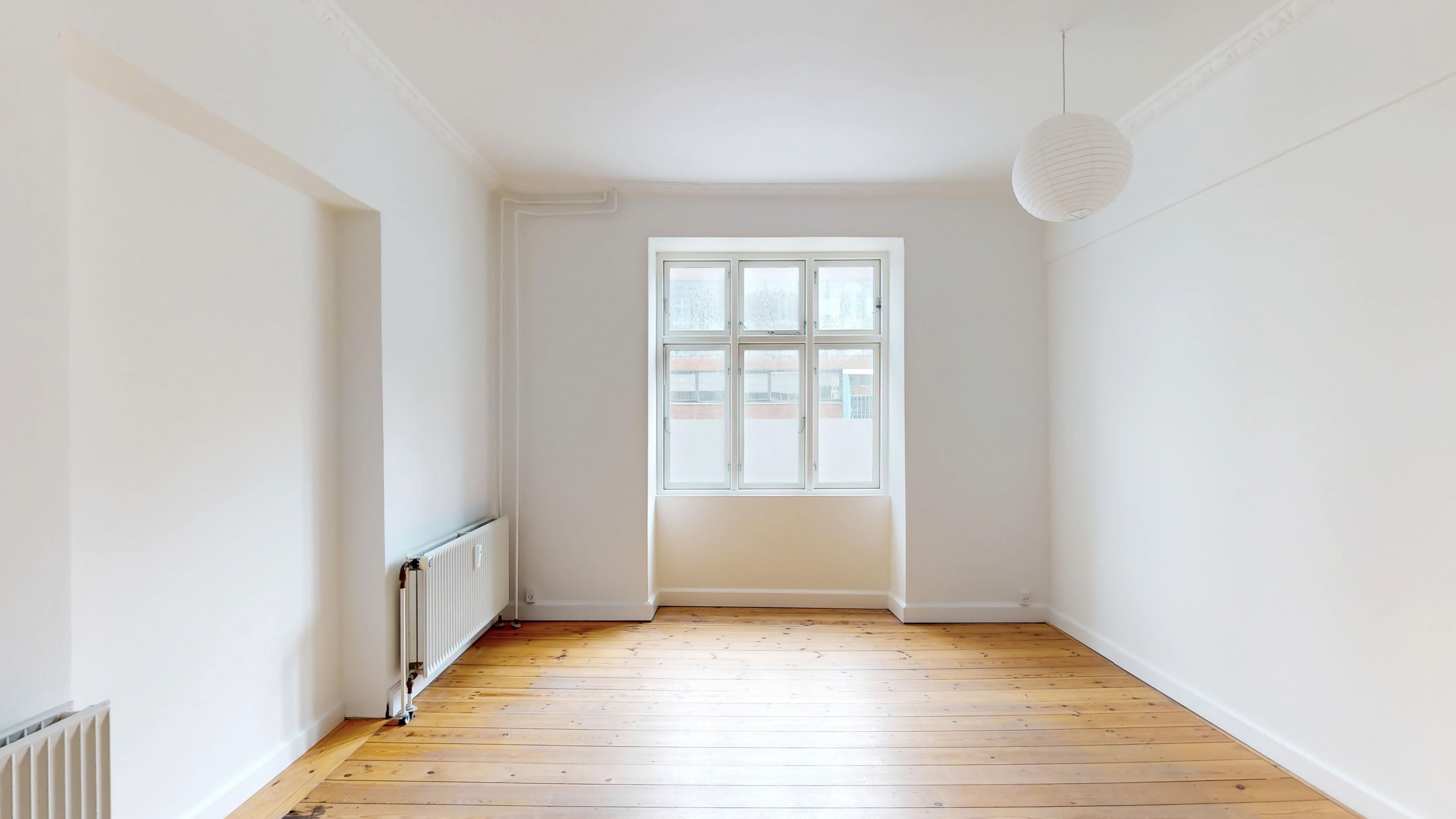 et tomt rum med højt loft og hårdt trægulv. Der er et vindue med hvid ramme og en hvid radiator på væggen. Loftet er hvidt og rummet har en hvidt indklædt væg.