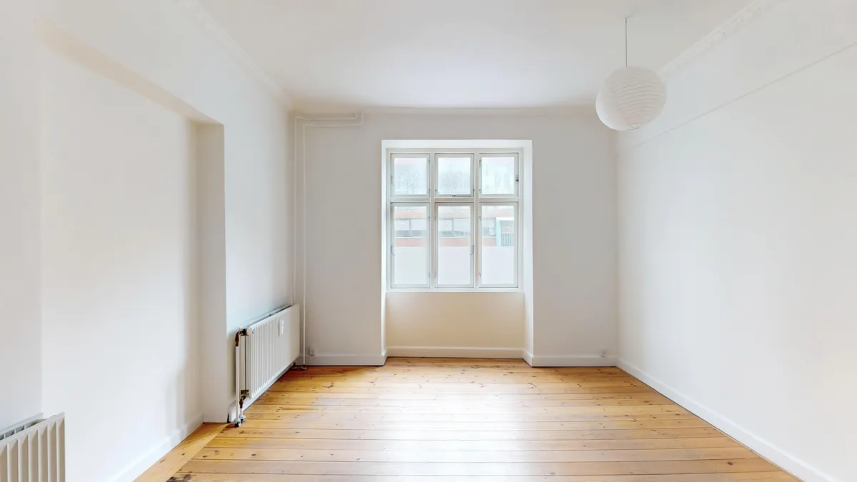 et tomt rum med højt loft og hårdt trægulv. Der er et vindue med hvid ramme og en hvid radiator på væggen. Loftet er hvidt og rummet har en hvidt indklædt væg.