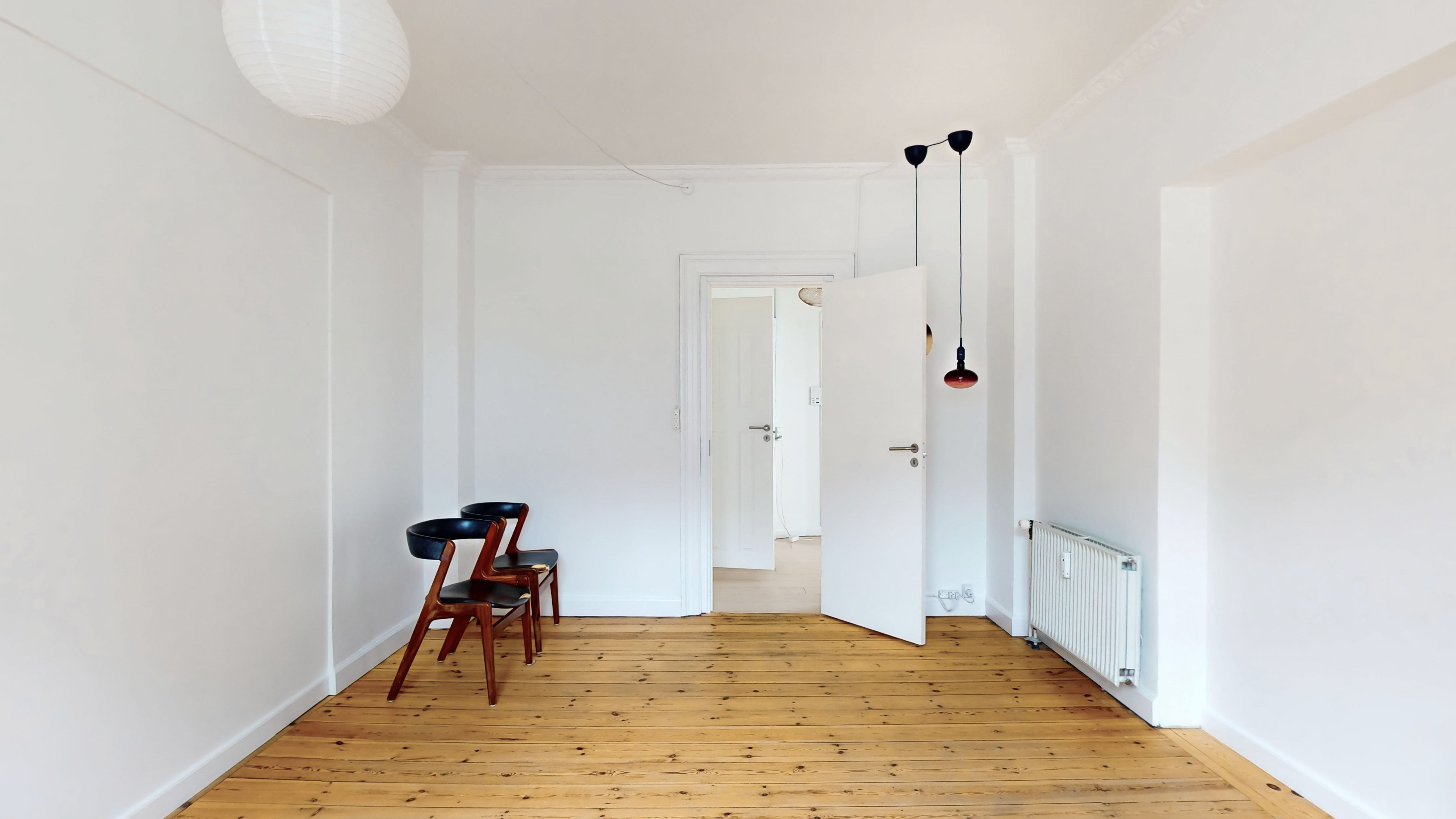 En tom værelse med hjørneplads og lyse lofter. To møbler med lyse træ og sorte sæder. En hvid radiator og to lyse pærekroner.