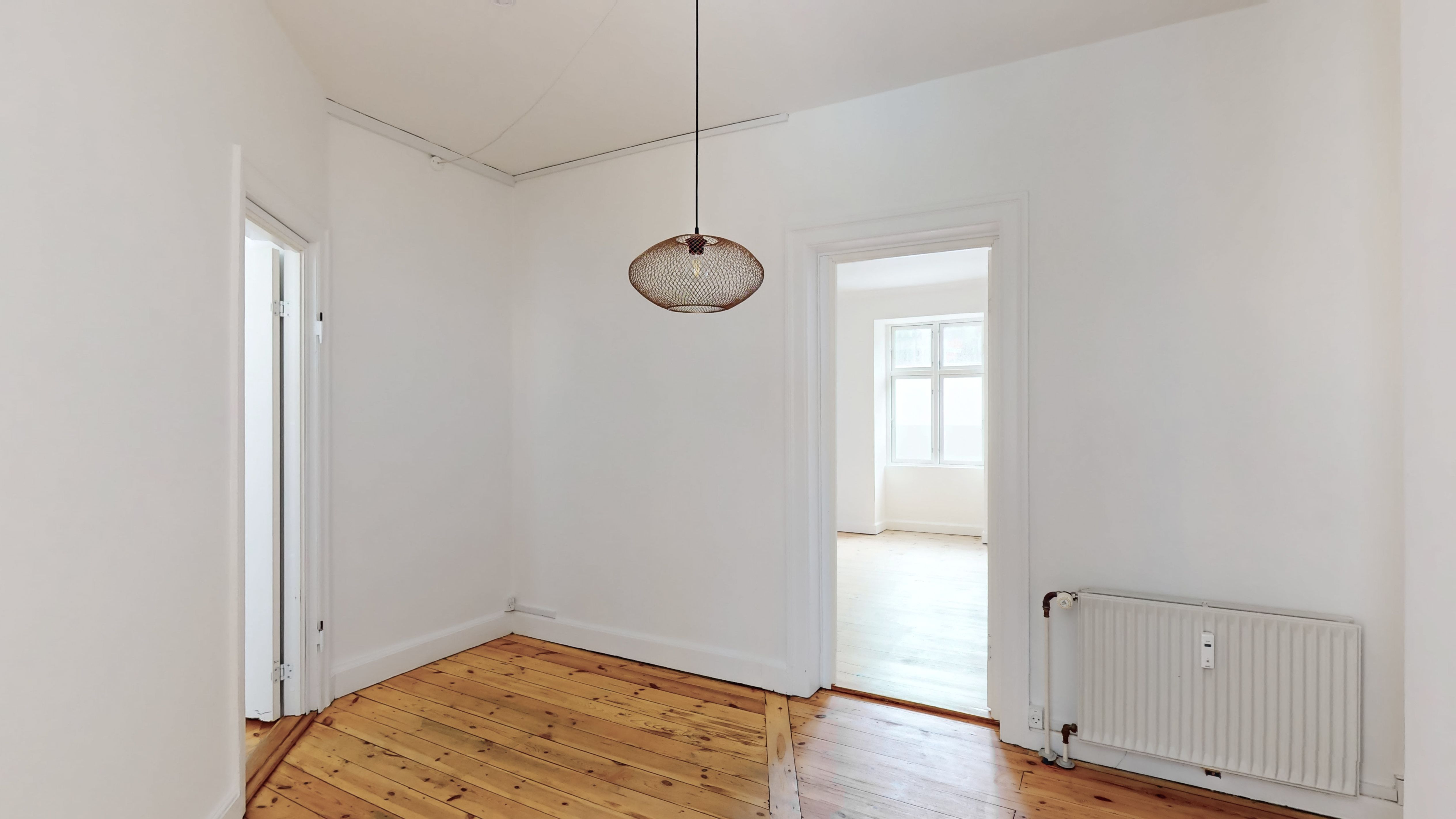 Kort gang med hjørnevinduet og åben dør. Hvid væg og trægulv. Rundt loftslampen i messing. Radiator og røre i hjørnet.