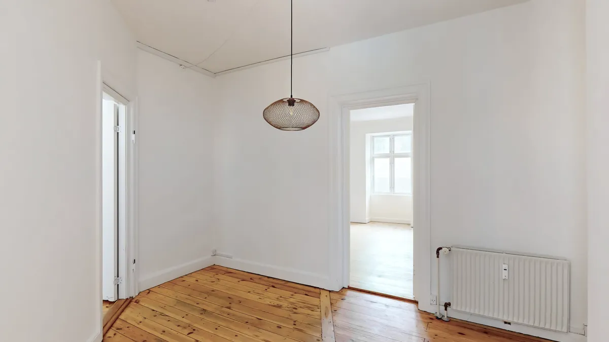 Kort gang med hjørnevinduet og åben dør. Hvid væg og trægulv. Rundt loftslampen i messing. Radiator og røre i hjørnet.