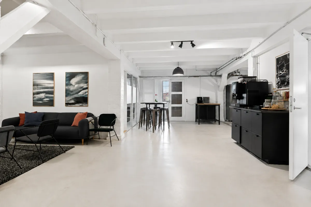 Kontor med højt til loftet, moderne møbler og lyse loftsbjælker i hvidt og stribet loft. Sovesofa med farverige puder og kunstværker i baggrunden.