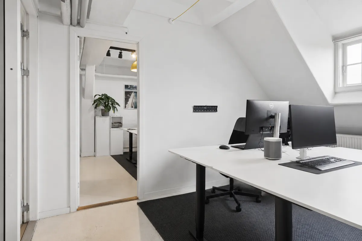 To monitorer og skrivebord med computer i lyse loftsrum med lysekron og plante i lyset. To lysegulve og hvide vægge.
