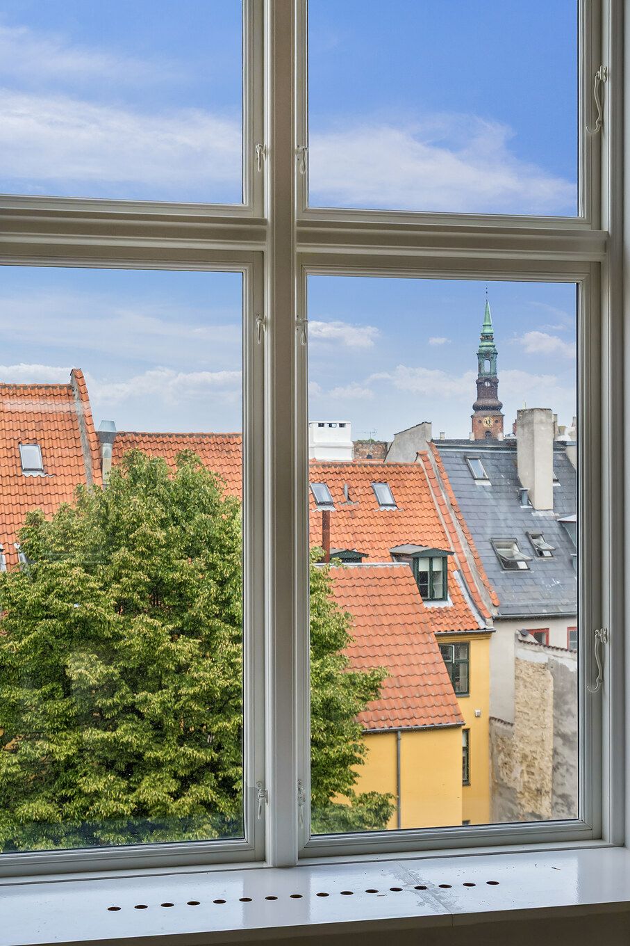 Lyst og charmerende kontor med udsigt over byens røde tage og tårne. Vinduet har hvide rammer og søjler, og der er en trætop i forgrunden. Solskinnet skinner på bygningerne og giver en varm atmosfære.