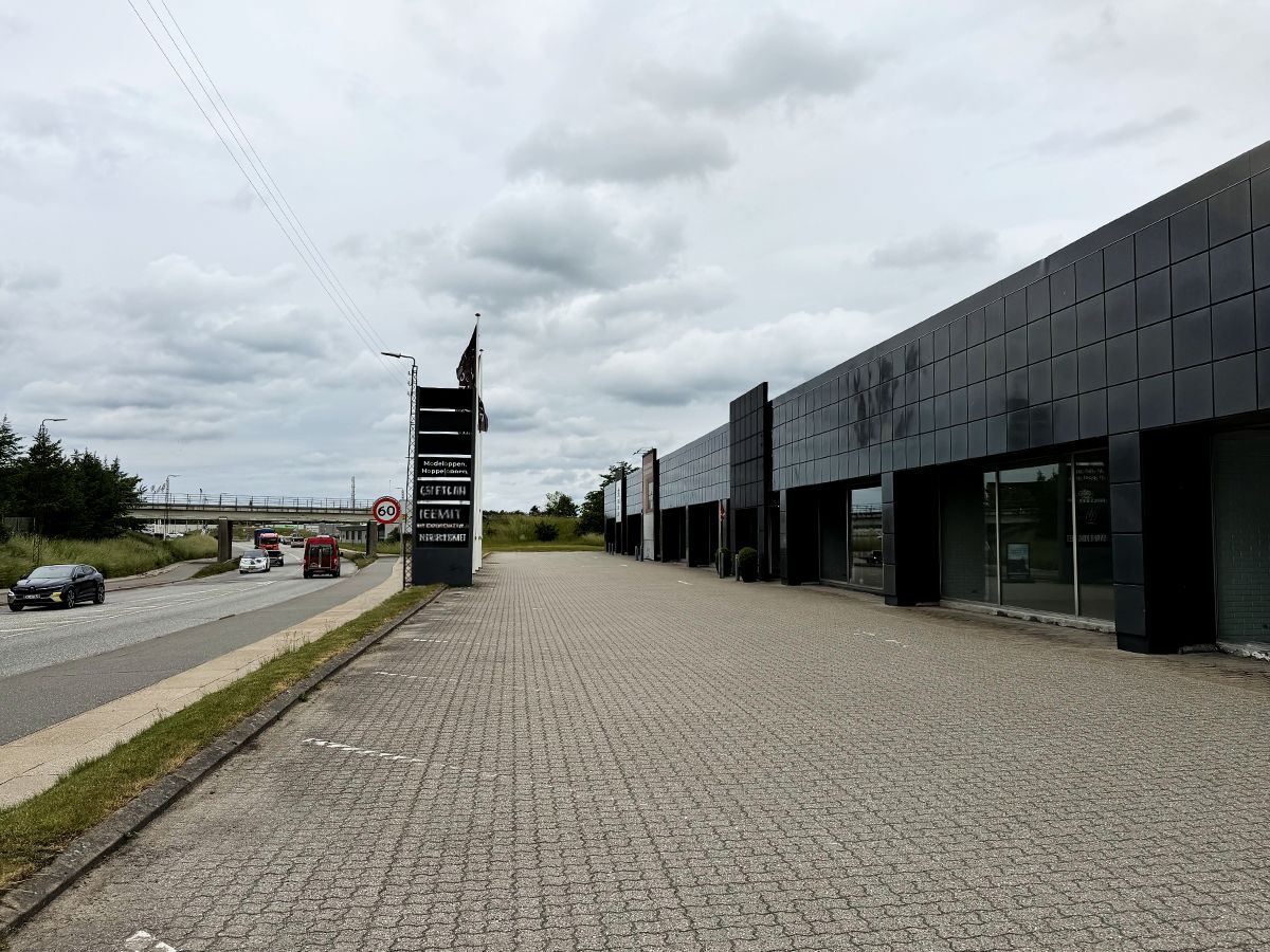Lejemålet til butik er på Farøvej 1C, 9800 Hjørring. Bygningen er i sort og hvid, og der er en stor parkeringsplads foran. En bil kører på hovedgaden