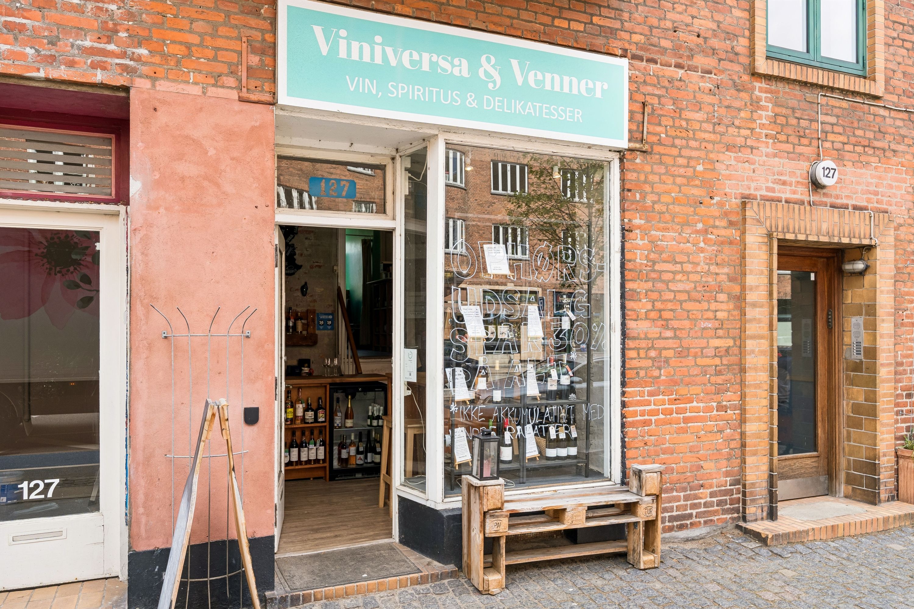 En butik med vin og alkohol med skiltet Viniversa og Venner. Der er flere vinflasker og andre drikkevarer i udsigten bag vinduet. Der er en træbænk og en lampe på fortauet.