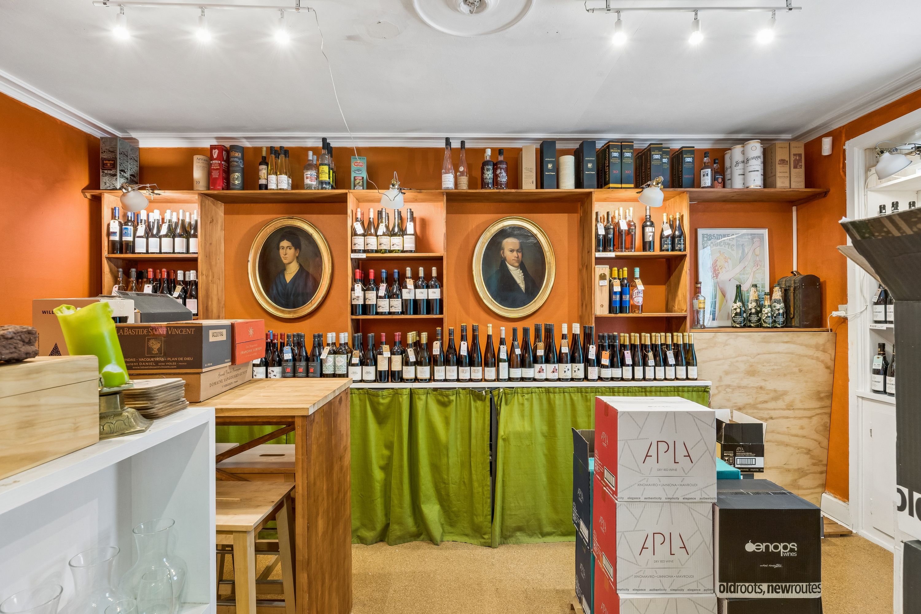 Store hylder med alkohol på butiksloftet. Flasker med etiketten APLÆ står i rækker. Fladskærmstv i loftet og møbler i gulv.