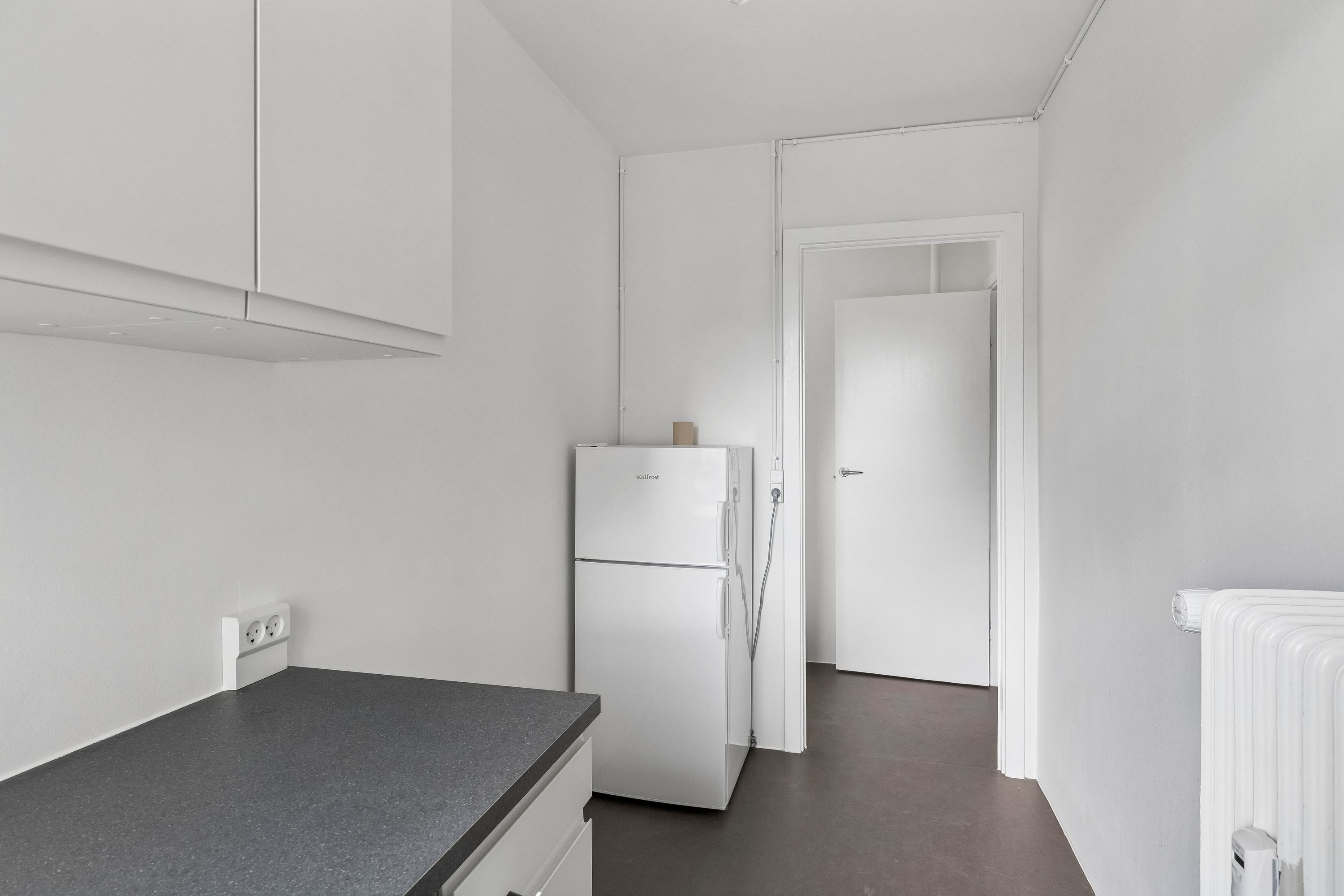 Lejlighed med hvidt køkken med en Væstfrost køleskab og krydsfinerbord. På væggen er der to hvide stikkontakter og en radiator samt hvide vægskabe