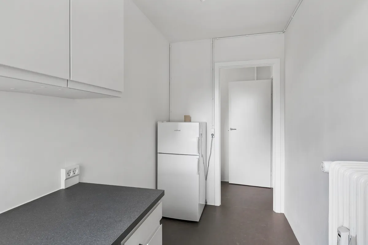 Lejlighed med hvidt køkken med en Væstfrost køleskab og krydsfinerbord. På væggen er der to hvide stikkontakter og en radiator samt hvide vægskabe
