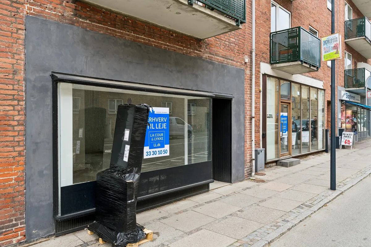 Butikstilbud på Ordrupvej 64, Charlottenlund. Lejetegnet er lavet af glas og metal. Stykkevis pakket møbel med sort emballage. På siden står der parkeringsskilt og salgsskilt.