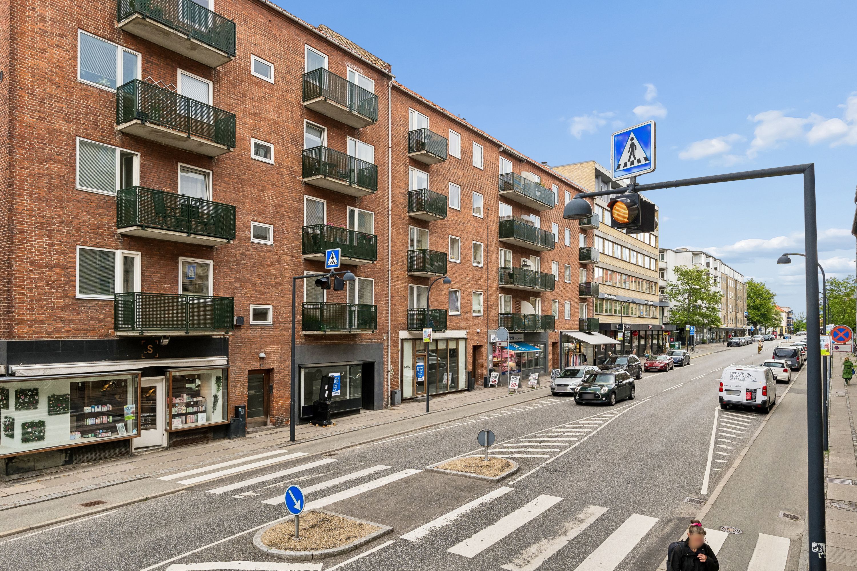 En tværgående gågade med større handelslokaler og parkerede biler og en person der krydser gaden på zebraovergangen