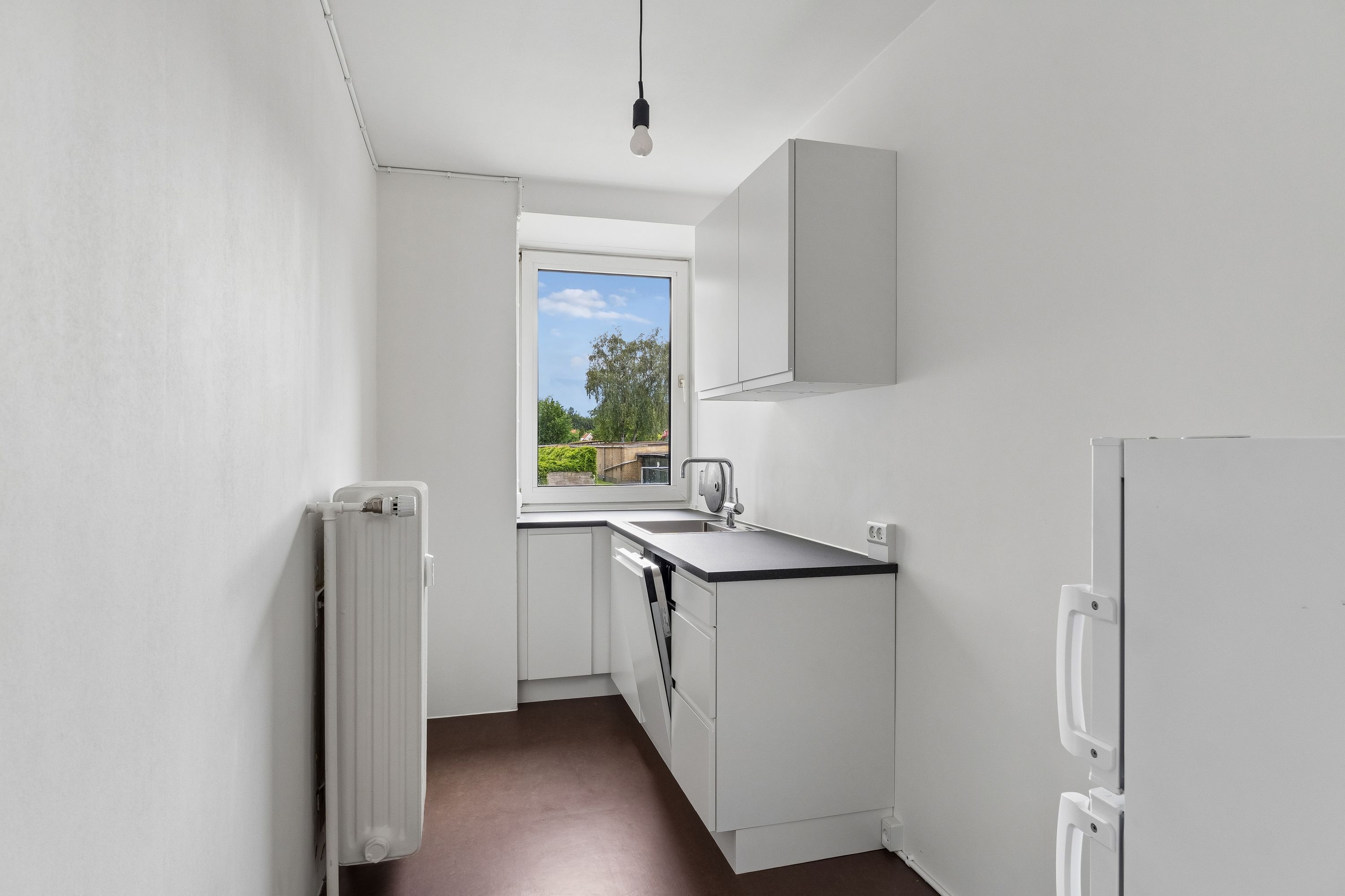 Hvidt køkken med hvide vægge og mørke gulve. Vindue med udsigt til grønne træer og hus. Hængende pærelampe. Hvidt køleskab med håndtag. Hvidt radiator og udtag.