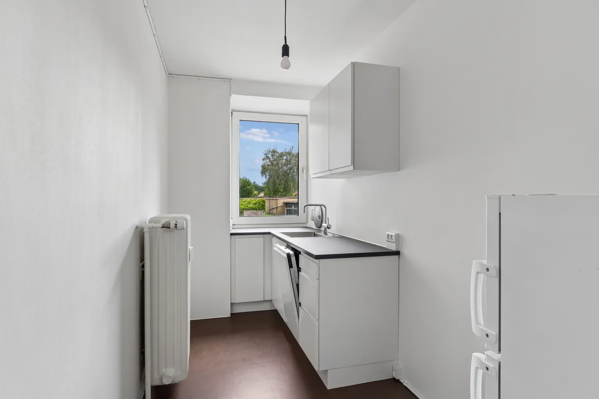Hvidt køkken med hvide vægge og mørke gulve. Vindue med udsigt til grønne træer og hus. Hængende pærelampe. Hvidt køleskab med håndtag. Hvidt radiator og udtag.