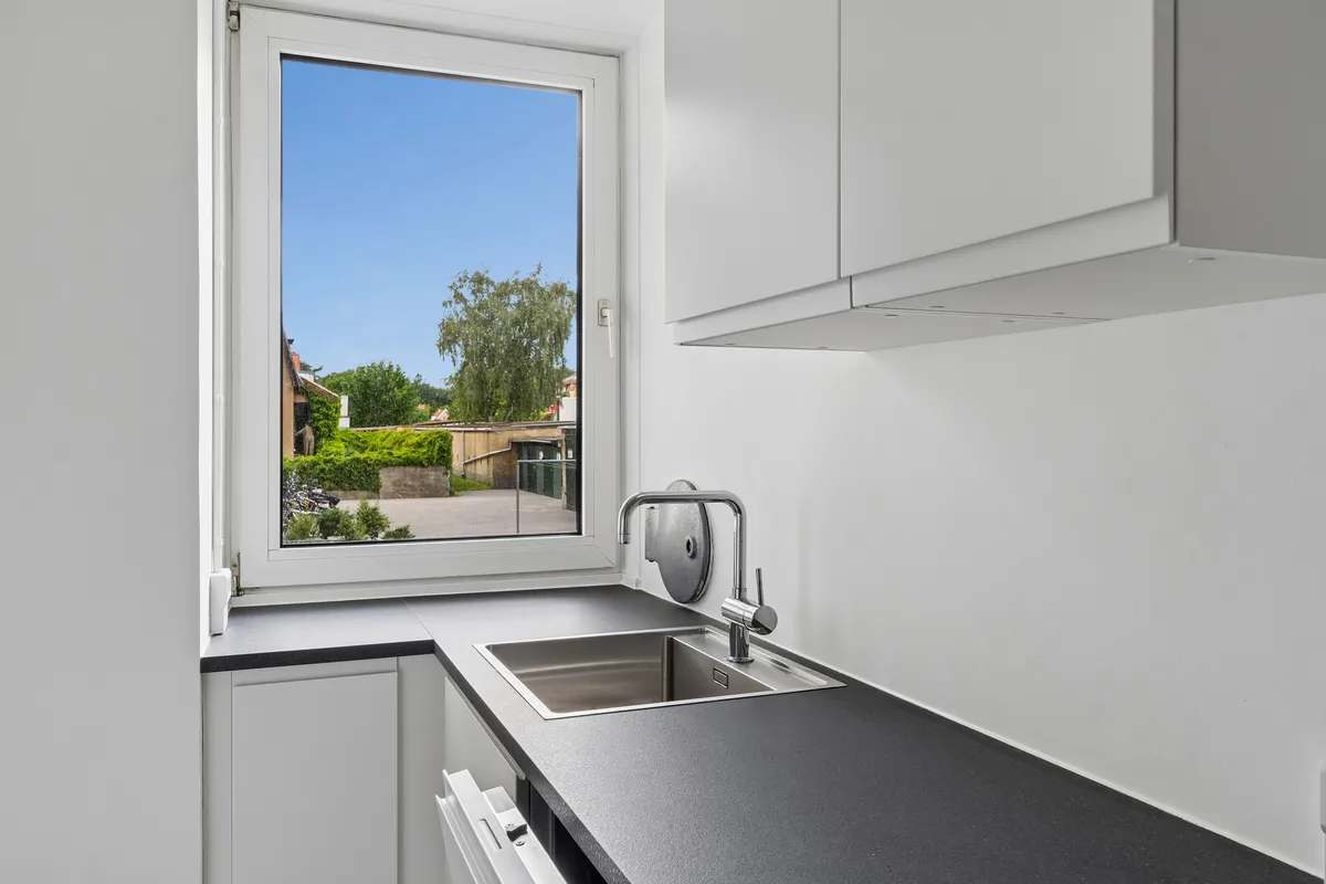 Moderne køkken med høj kvalitet og lyse farver, hvidt loft, hvidt gulv og sort granitkøkkenbord med rustfrit stålvandhane og et stort glasvindue med udsigt til grønne haver