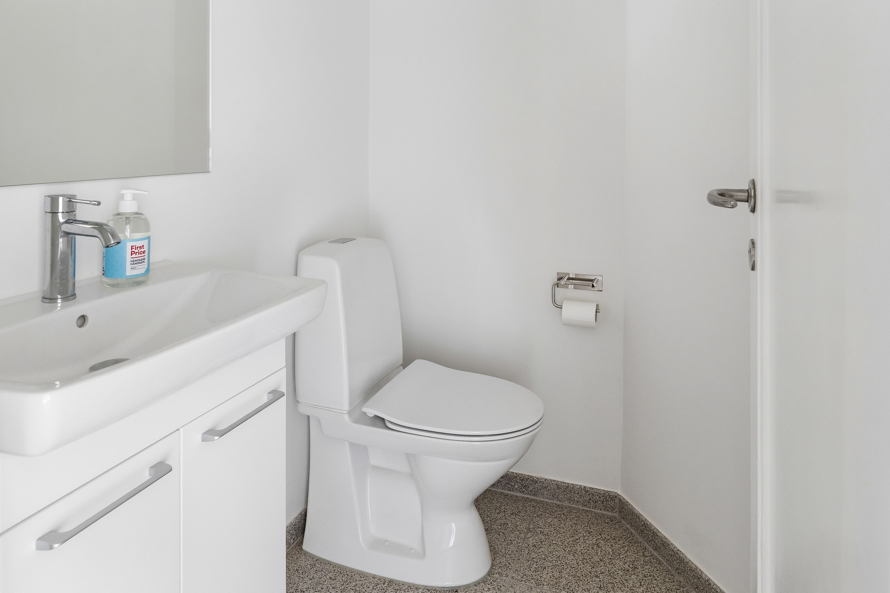 Et moderne hvidt badeværelse med et toilet, et hvidt vasketøjsskab, et hvidt spejl og en hvid vask med håndsæbe.