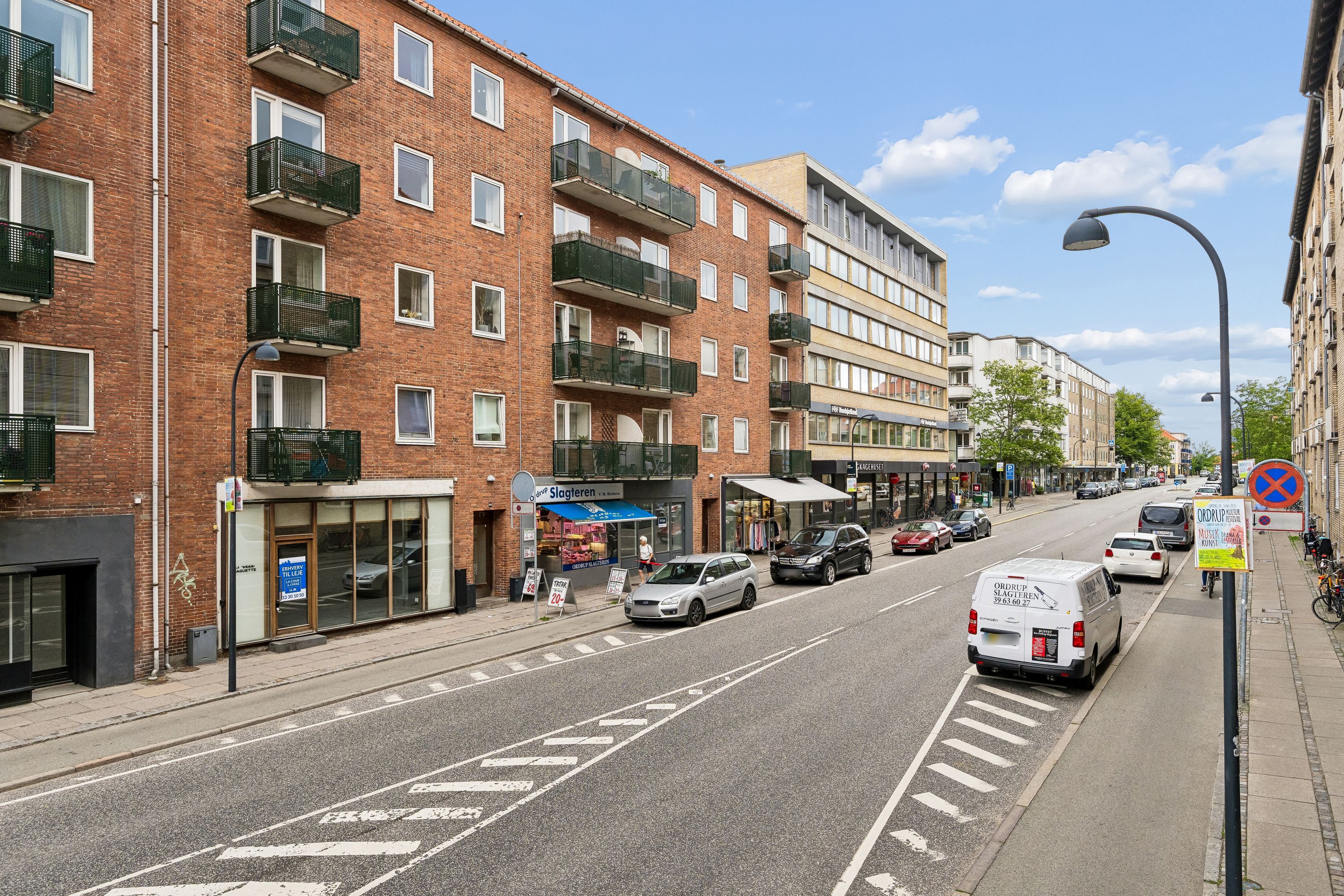 En række butikker med glasfronter og balkoner på Ordrupvej 62B. En forretning med navnet Slagleren og flere biler parkeret på gaden og på siderne