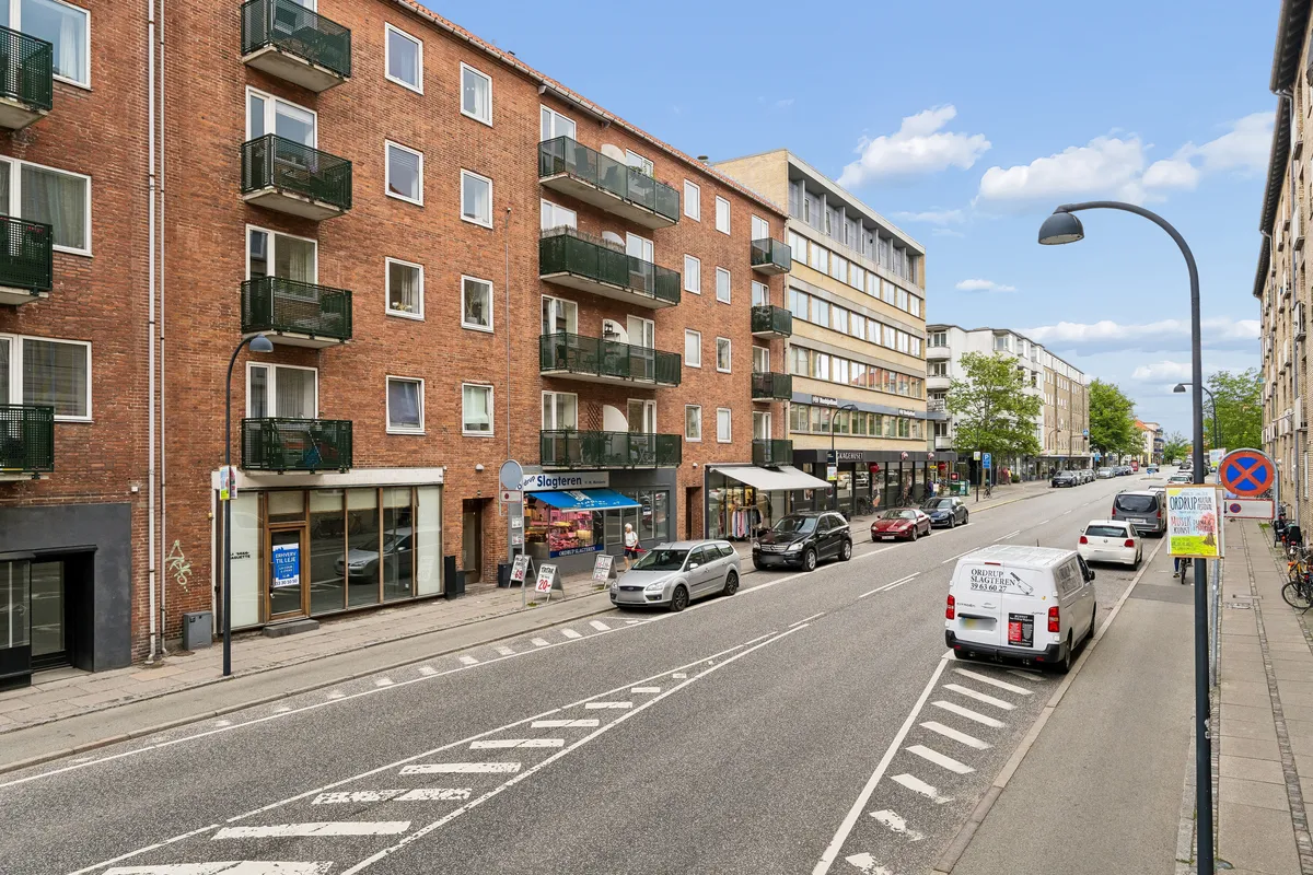 En række butikker med glasfronter og balkoner på Ordrupvej 62B. En forretning med navnet Slagleren og flere biler parkeret på gaden og på siderne