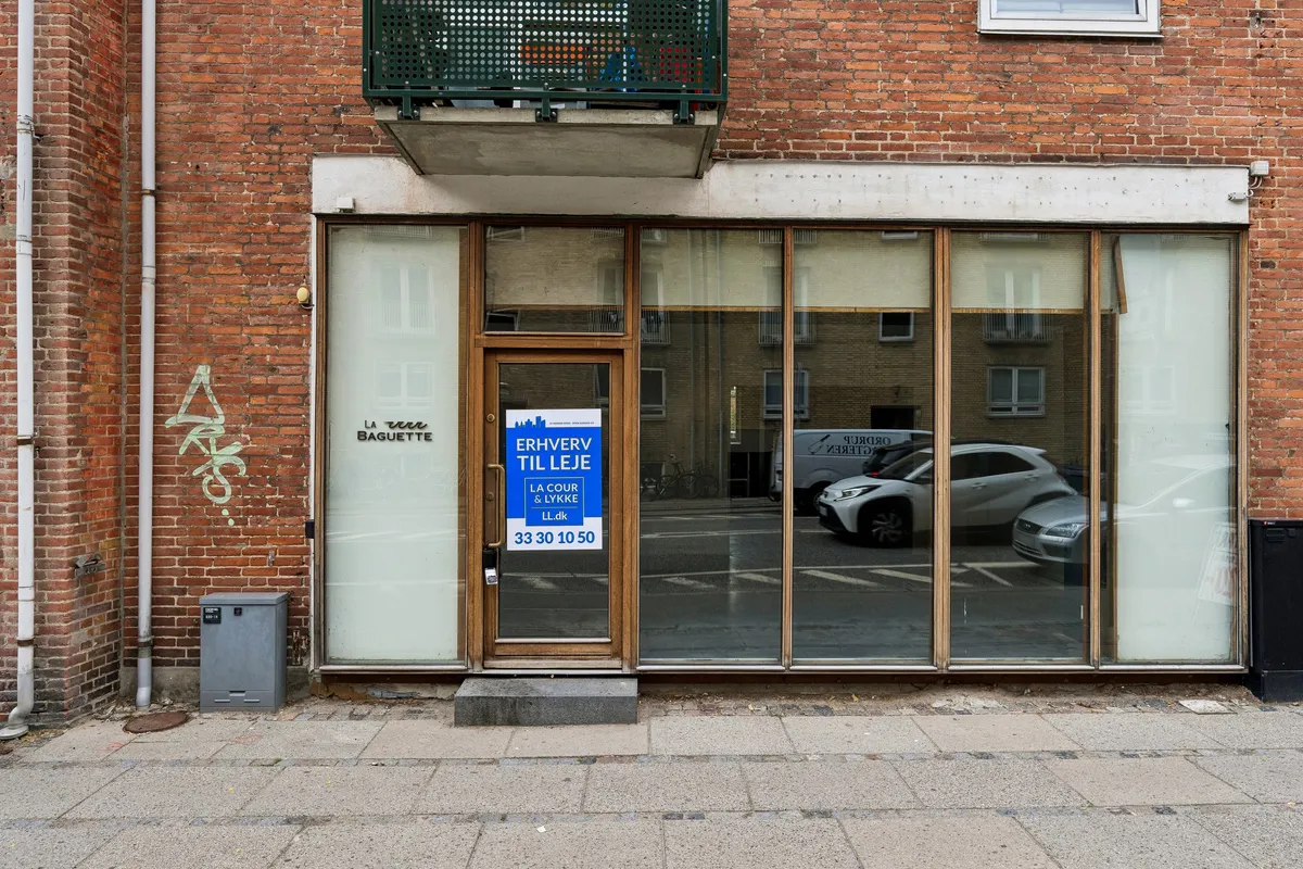 Lejemål i Charlottenlund. Butiksfront med glasdøre og vindue. Balkon og graffiti på rødt murstensvæg. Parkerede biler og bygning i udsigt.